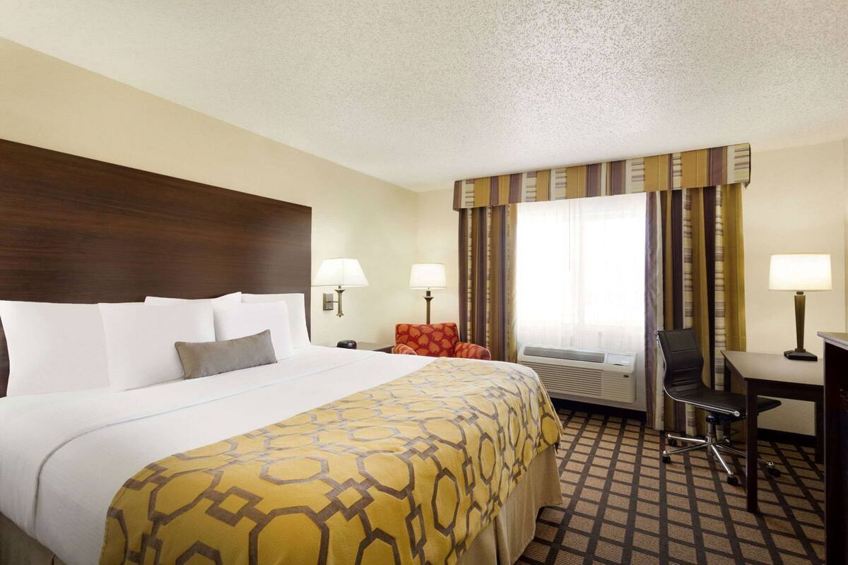 A hotel room with a large bed, two bedside lamps, a desk with a chair, and an air conditioning unit.