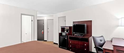 A hotel room with a large bed, a desk with a chair, a television, a minibar, and a door leading to another room.
