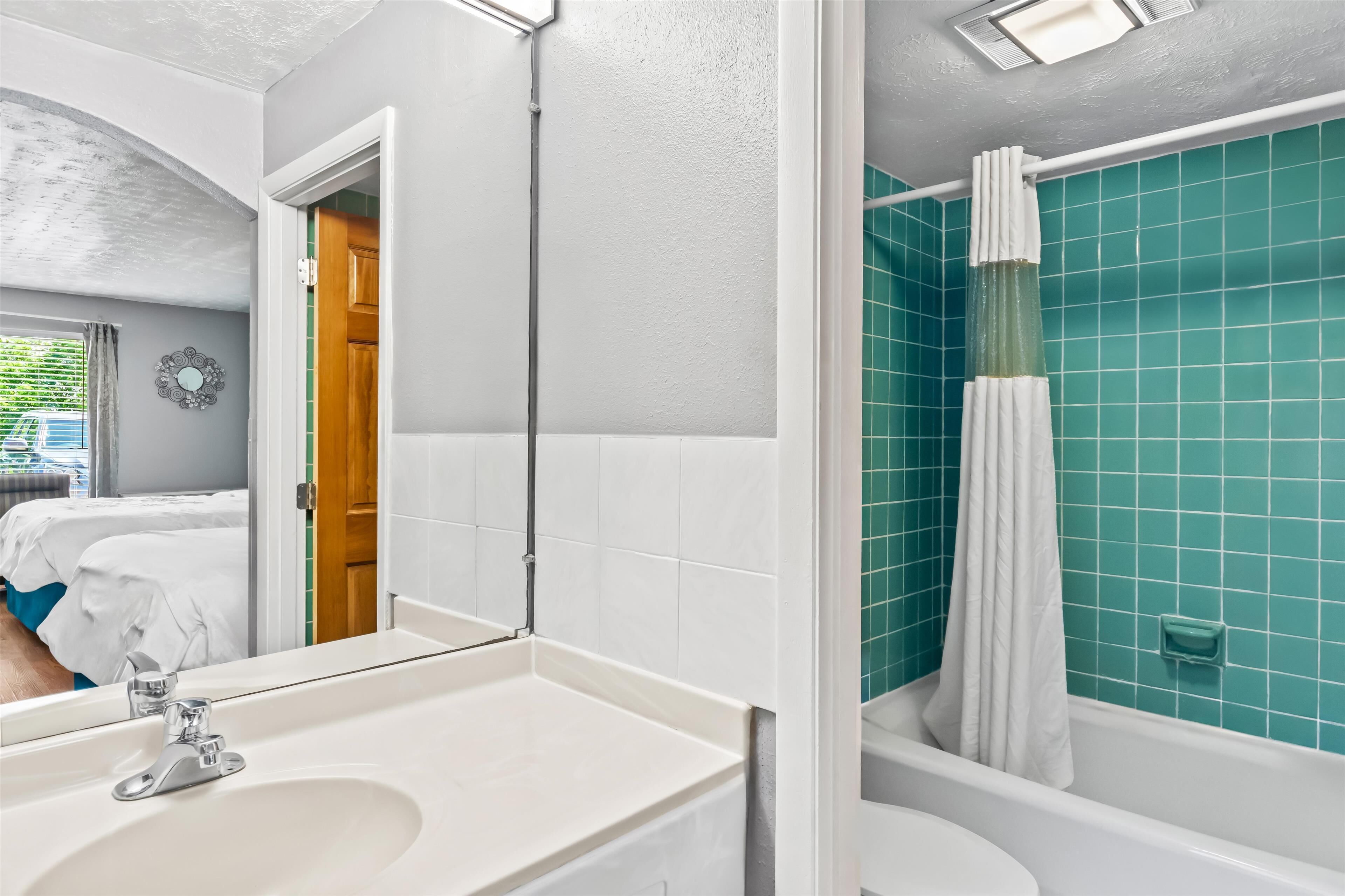 A bathroom with a white sink, a mirror, a shower with a white curtain, and teal tiled walls.