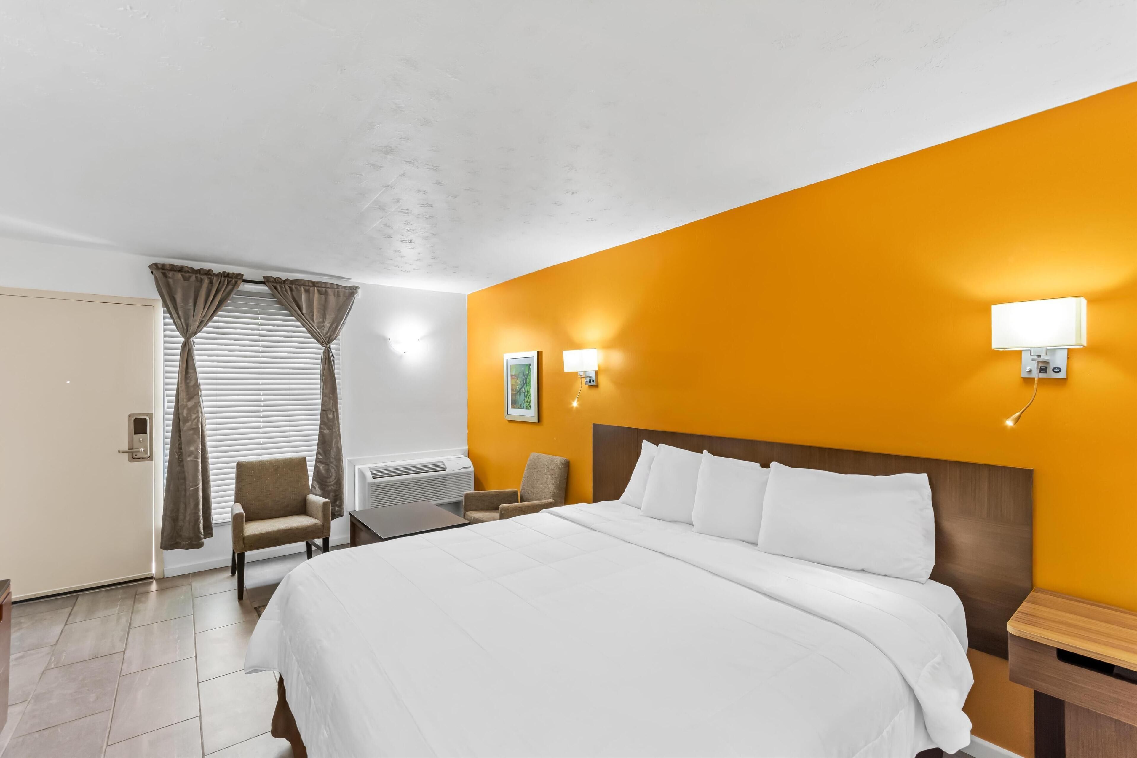 Habitación de hotel con una cama grande, un escritorio, dos sillas, una ventana con cortinas y una lámpara fijada a la pared.