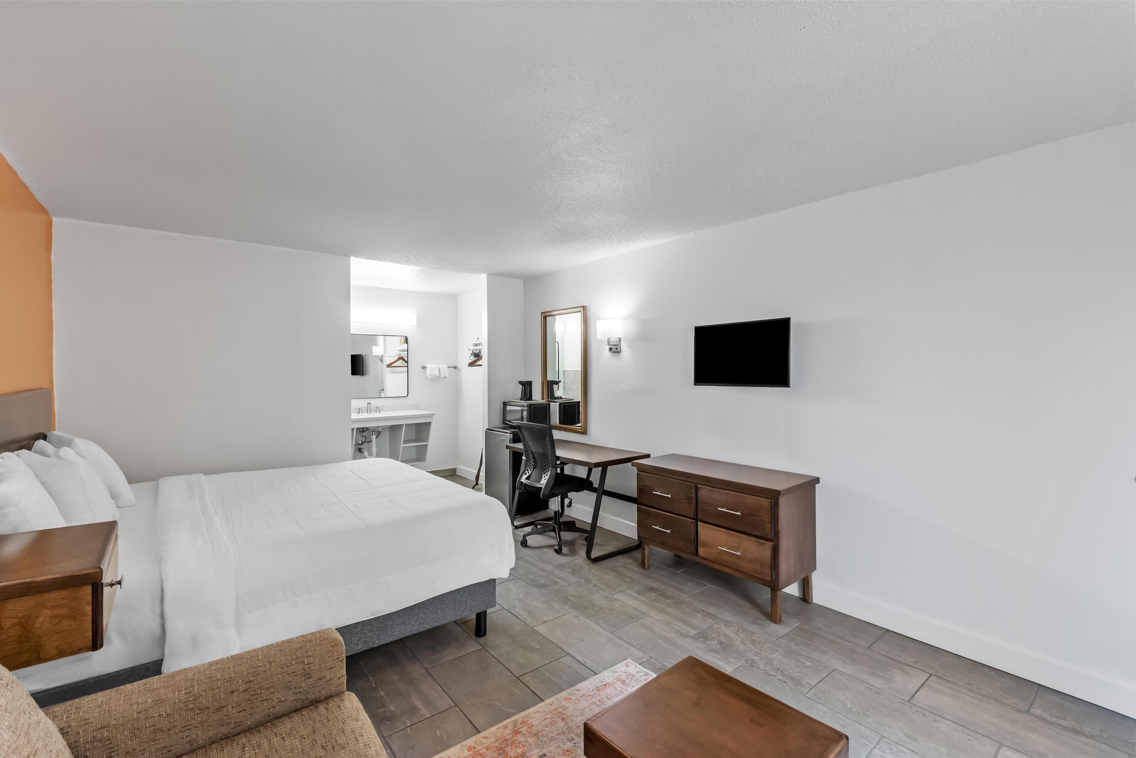 A hotel room with a bed, a desk, a chair, a dresser, a TV, and a bathroom visible in the background.