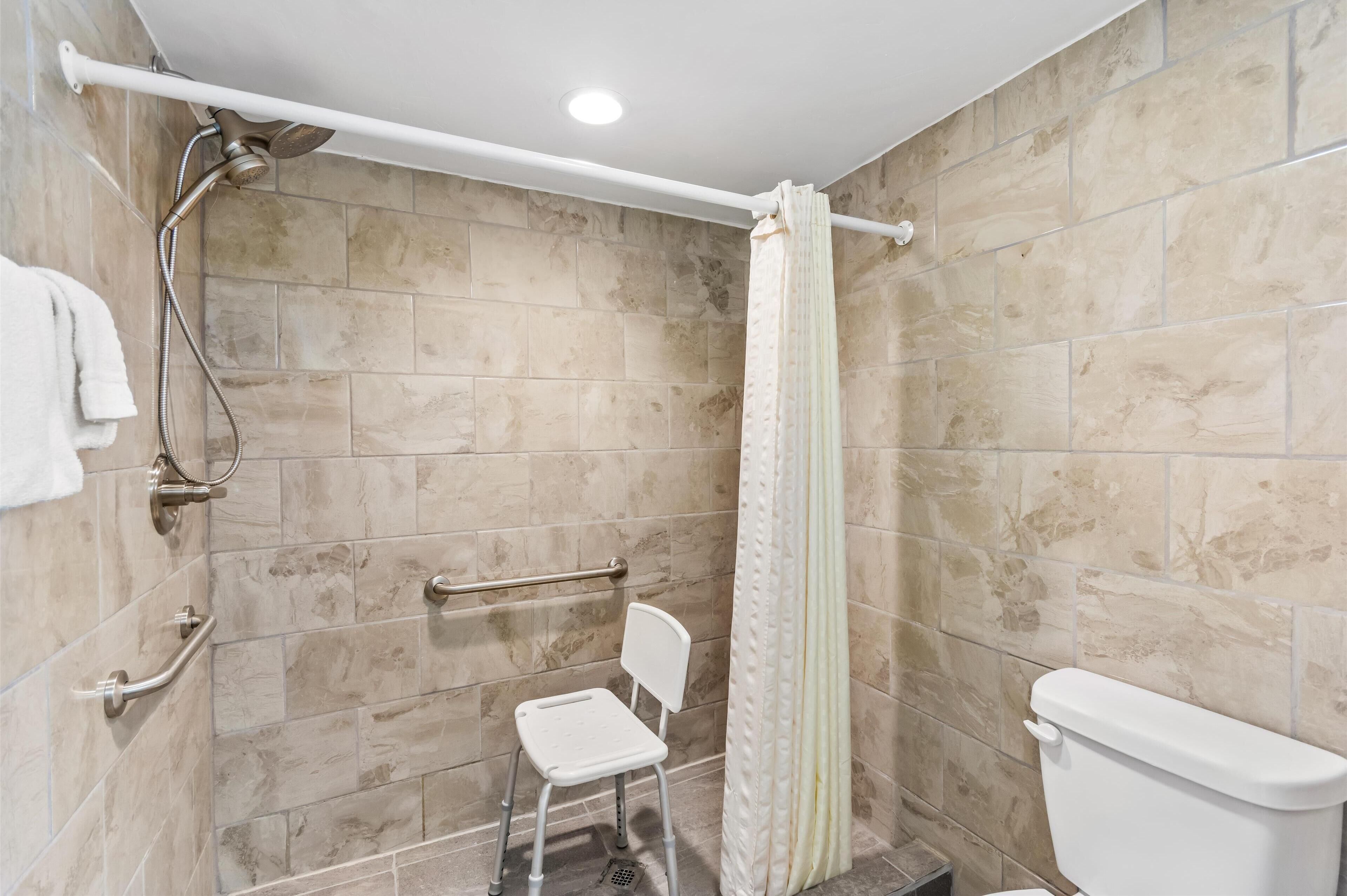 A bathroom with beige tiles, a white toilet, a shower with a white curtain, a grab bar, and a white chair.
