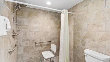 A bathroom with beige tiles, a white toilet, a shower with a white curtain, a grab bar, and a white chair.