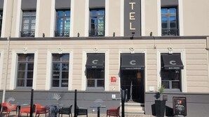 Une façade d’hôtel avec une enseigne indiquant « HÔTEL » au-dessus de l’entrée. On y trouve des espaces extérieurs aménagés avec des tables et des chaises.
