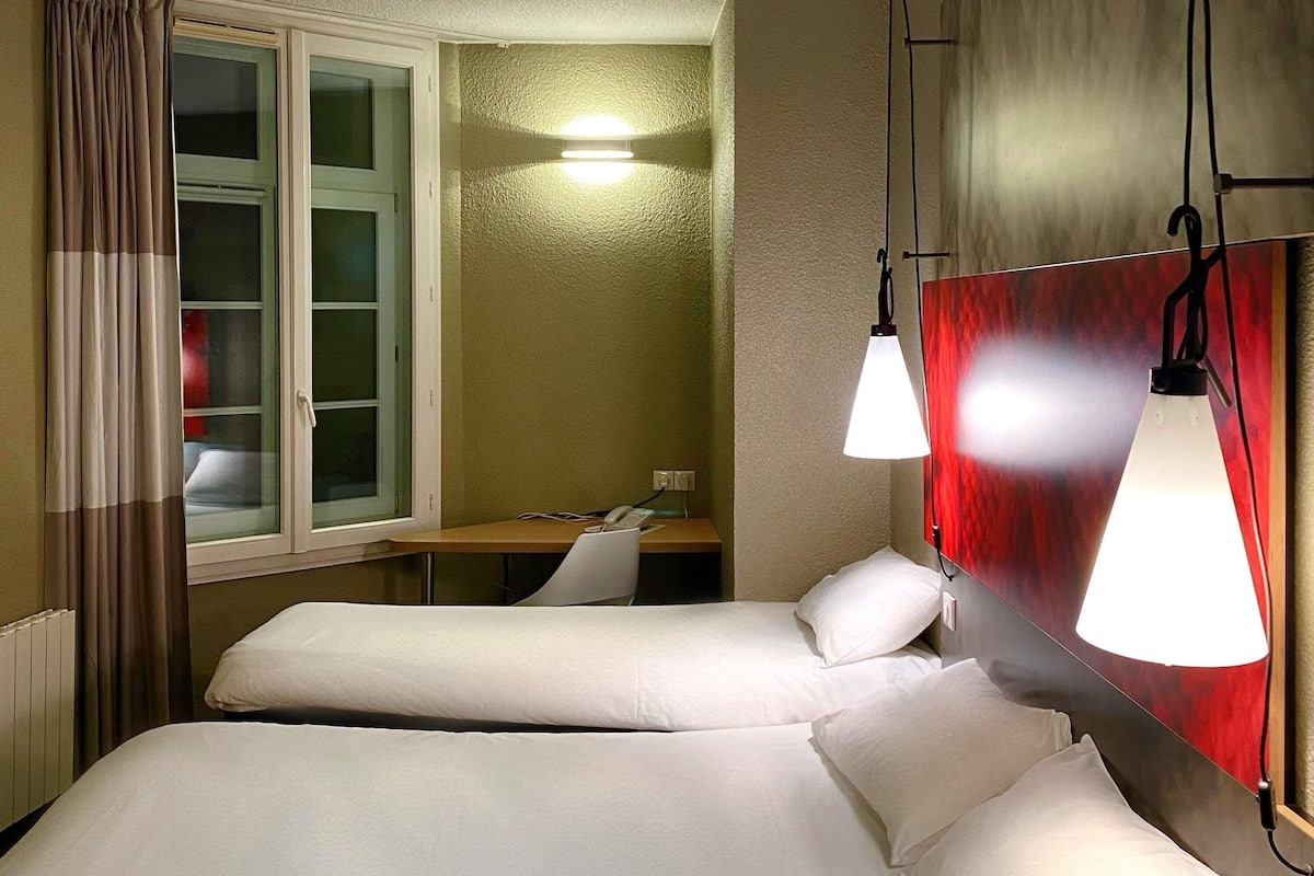 A hotel room with two beds, a desk, a window with curtains, and a wall-mounted light fixture.