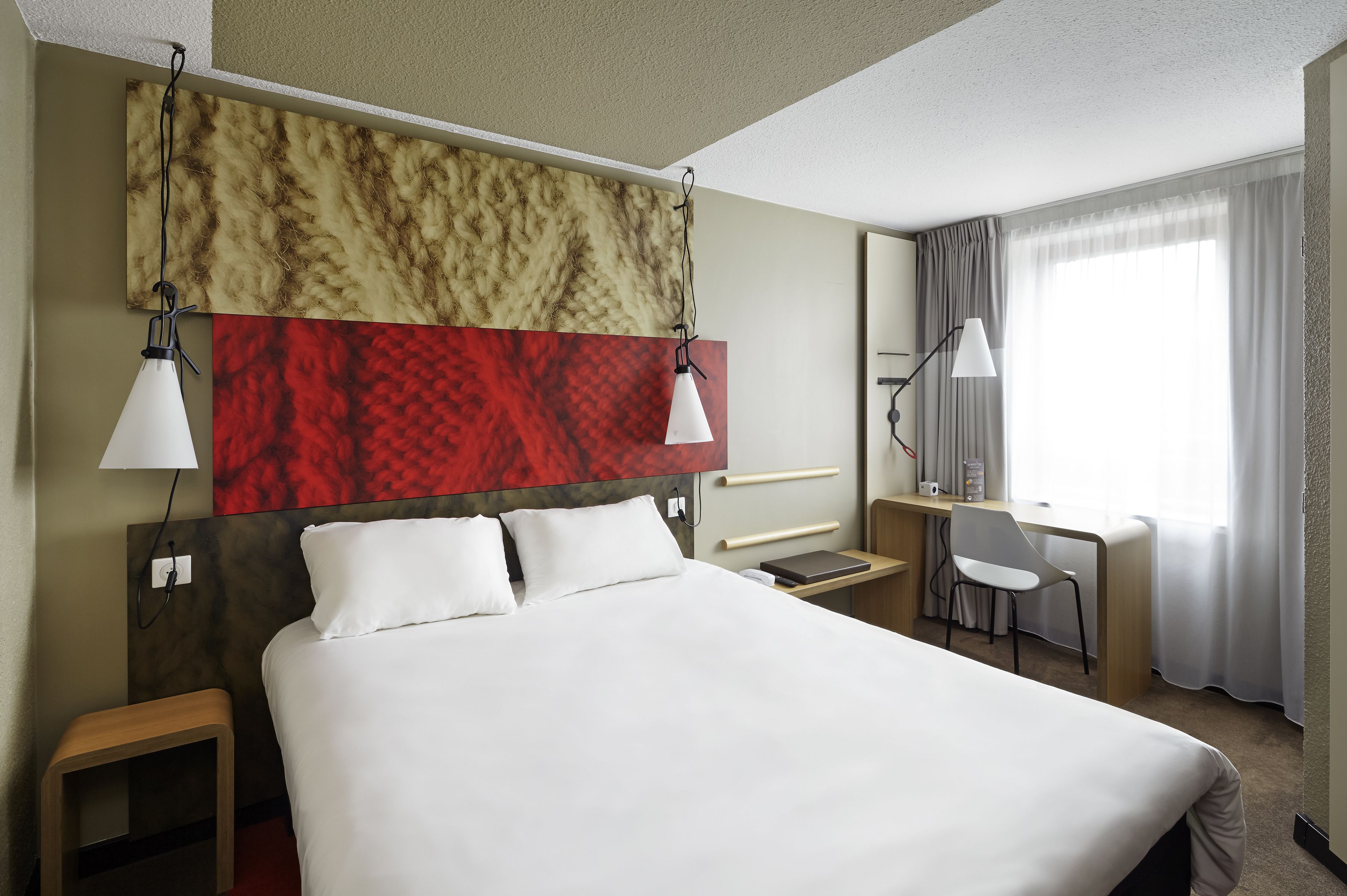Una habitación de hotel con una cama grande, dos mesitas de noche, un escritorio con silla, una ventana con cortinas y dos lámparas fijadas a la pared.