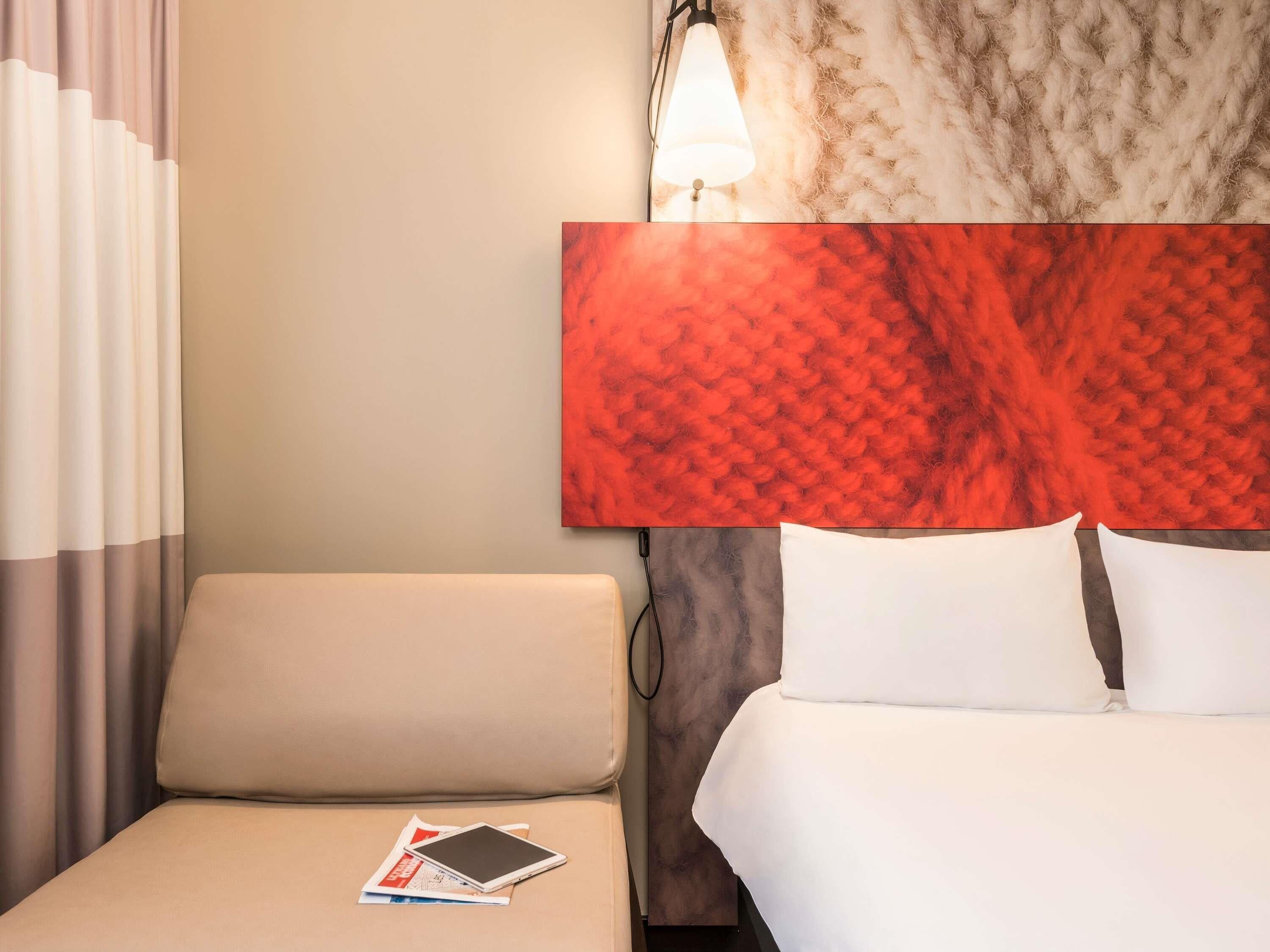 Een hotelkamer met een bed, een nachtkastje met een tablet, een lamp en een groot abstract schilderij aan de muur.