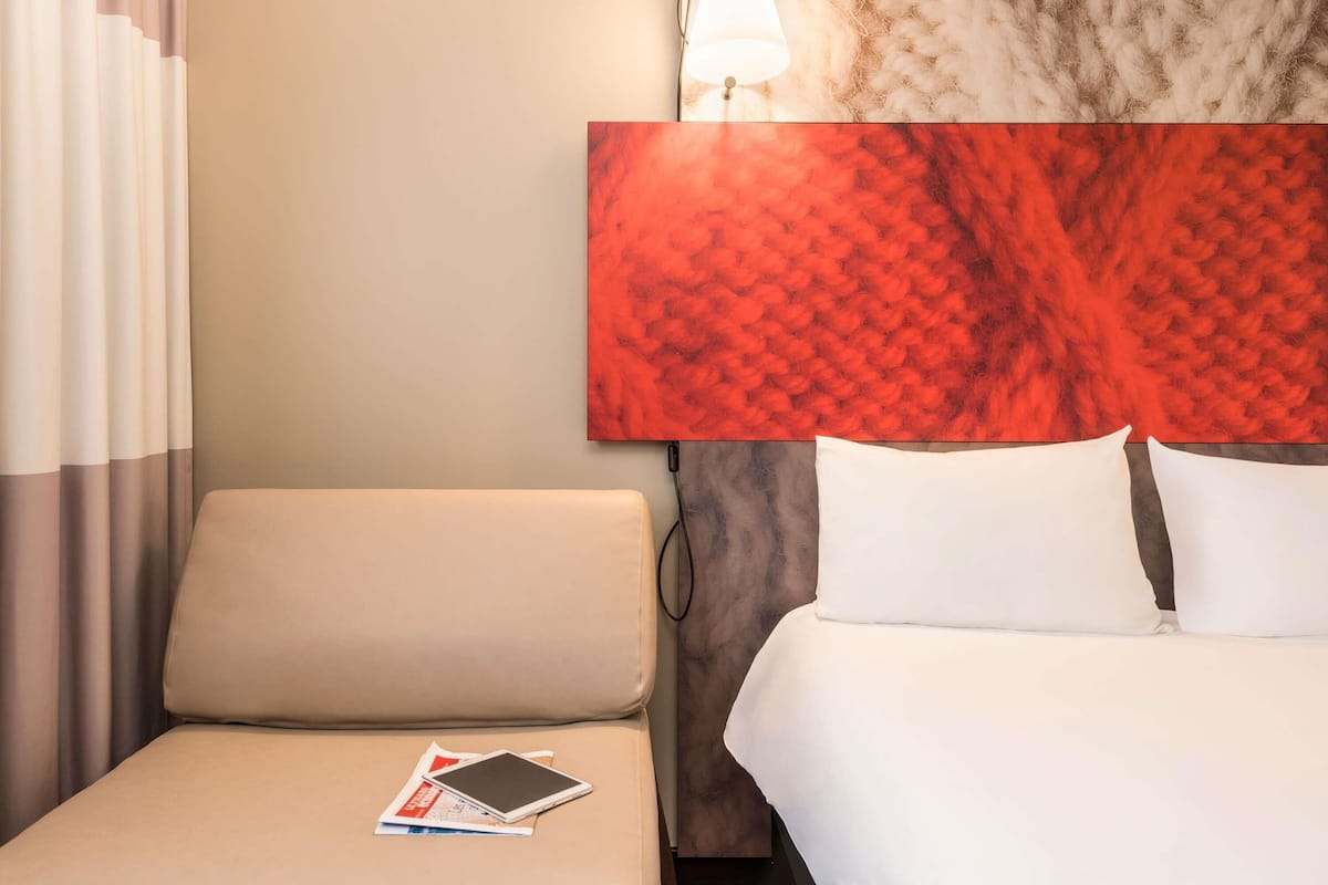 Habitación de hotel con cama, mesita de noche con una tablet, lámpara y un cuadro abstracto grande en la pared.