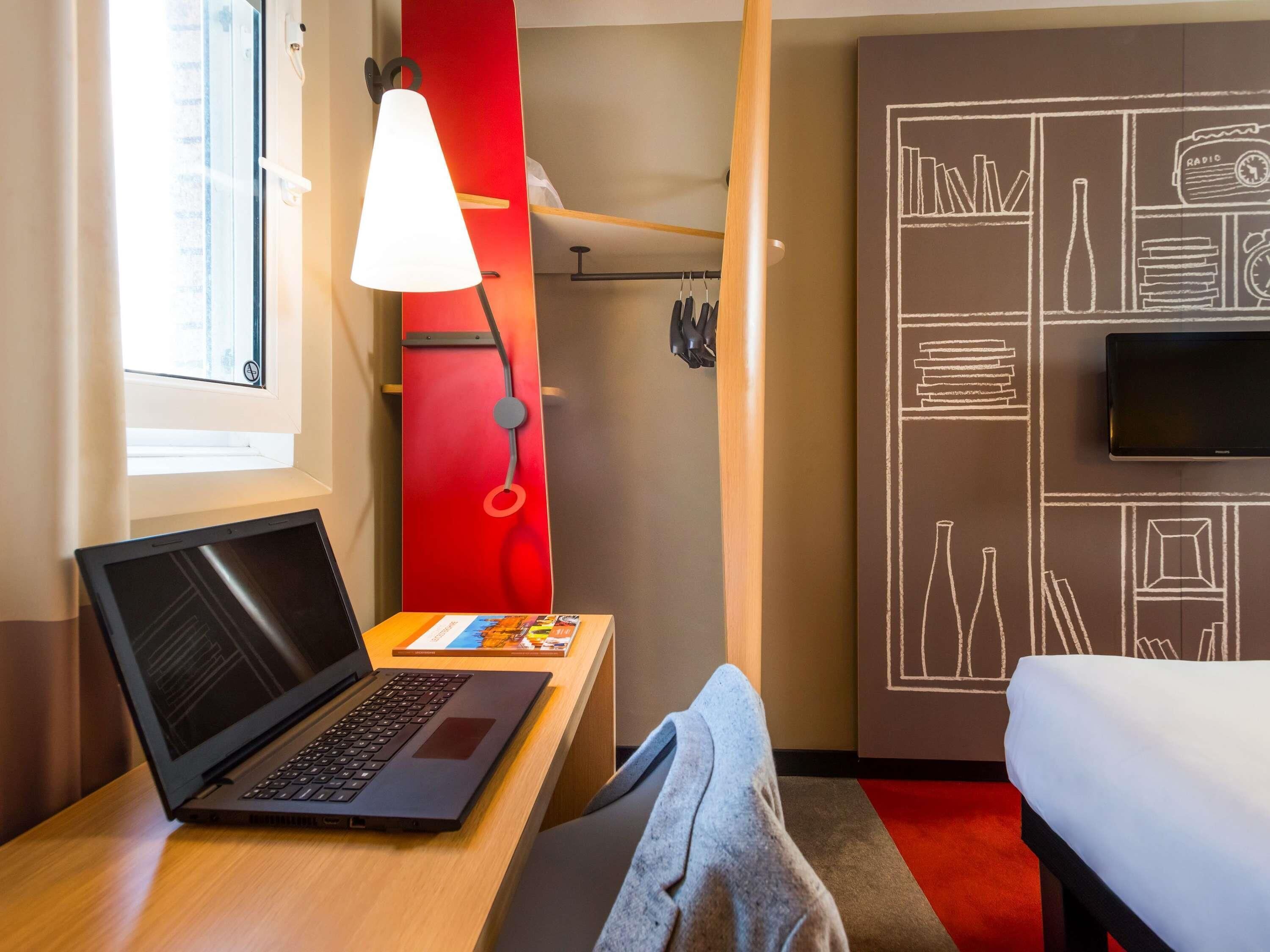 A hotel room with a laptop on a desk, a bed, a TV, and a wall with a chalkboard design.