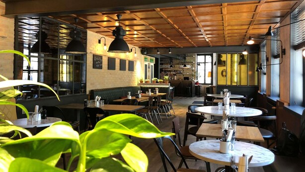 L’intérieur d’un restaurant avec des poutres en bois au plafond, des tables rondes et des plantes vertes.