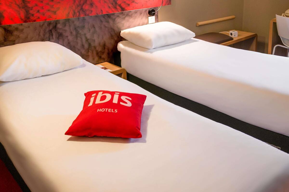 Habitación de hotel con dos camas, almohada roja con el texto 'ibis HOTELS', una mesita de noche de madera y una pared roja.