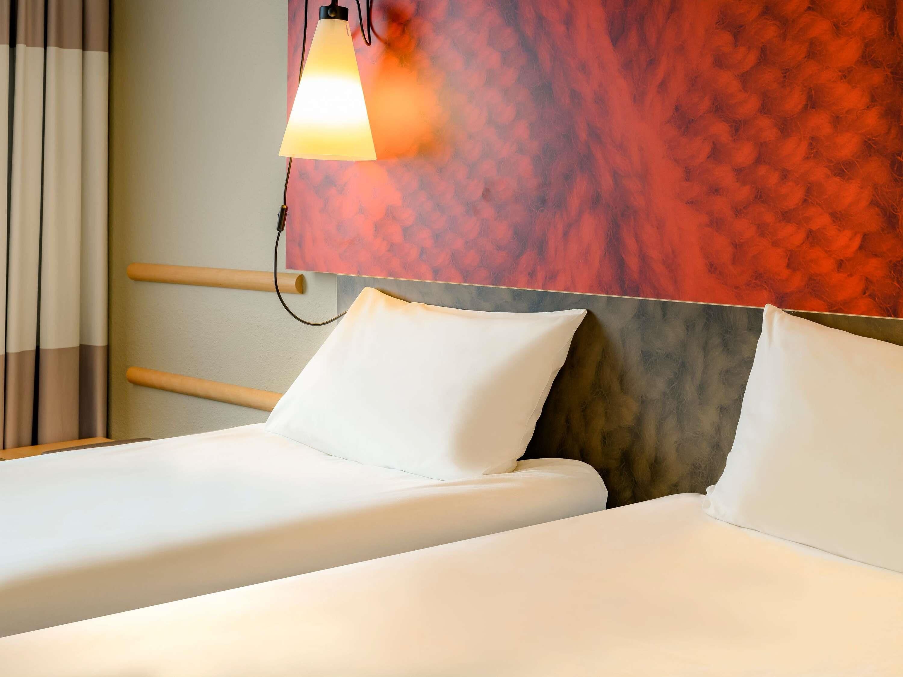 A hotel room with a bed, white bedding, a headboard with a textured wall behind it, and a wall-mounted lamp.