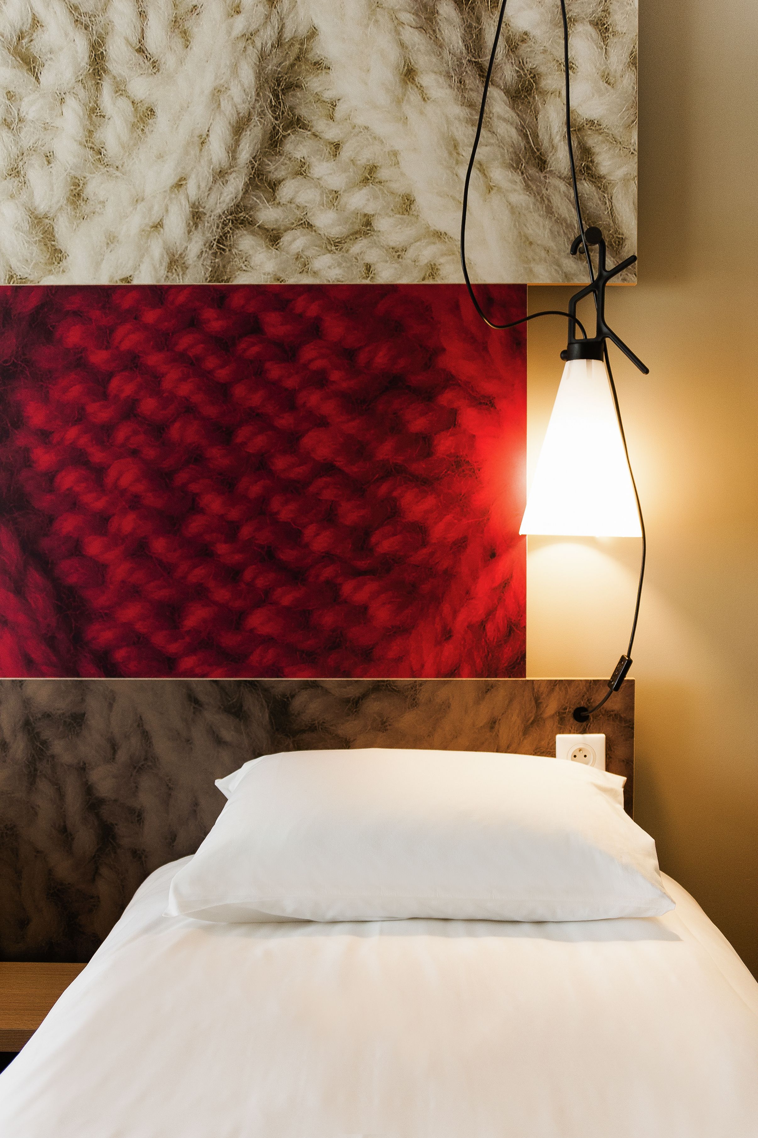 Una cama con ropa de cama blanca, una pared texturizada de color rojo y una lámpara fijada a la pared.