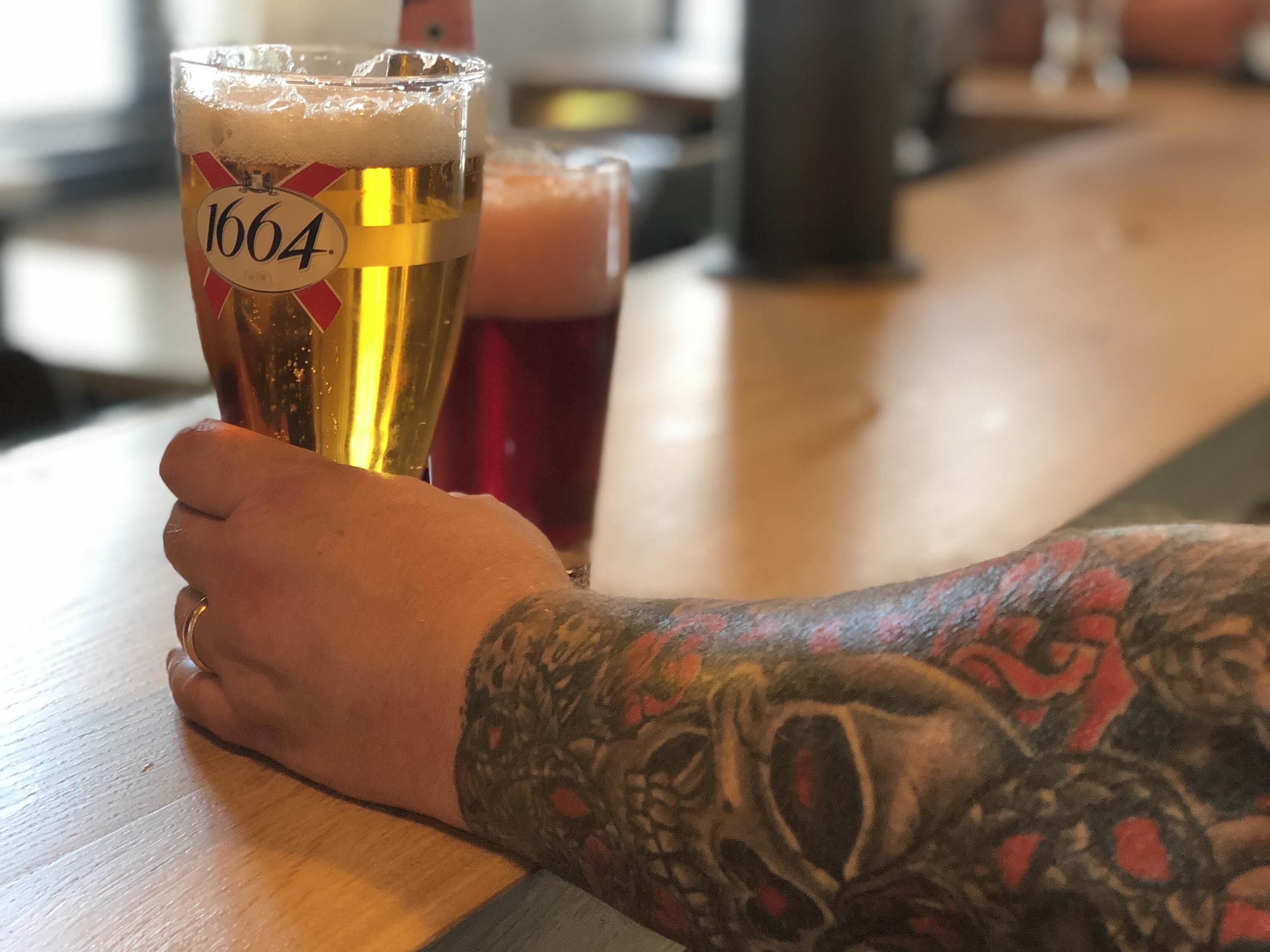 A person holding a glass of beer with a label reading "1664" on a wooden table.
