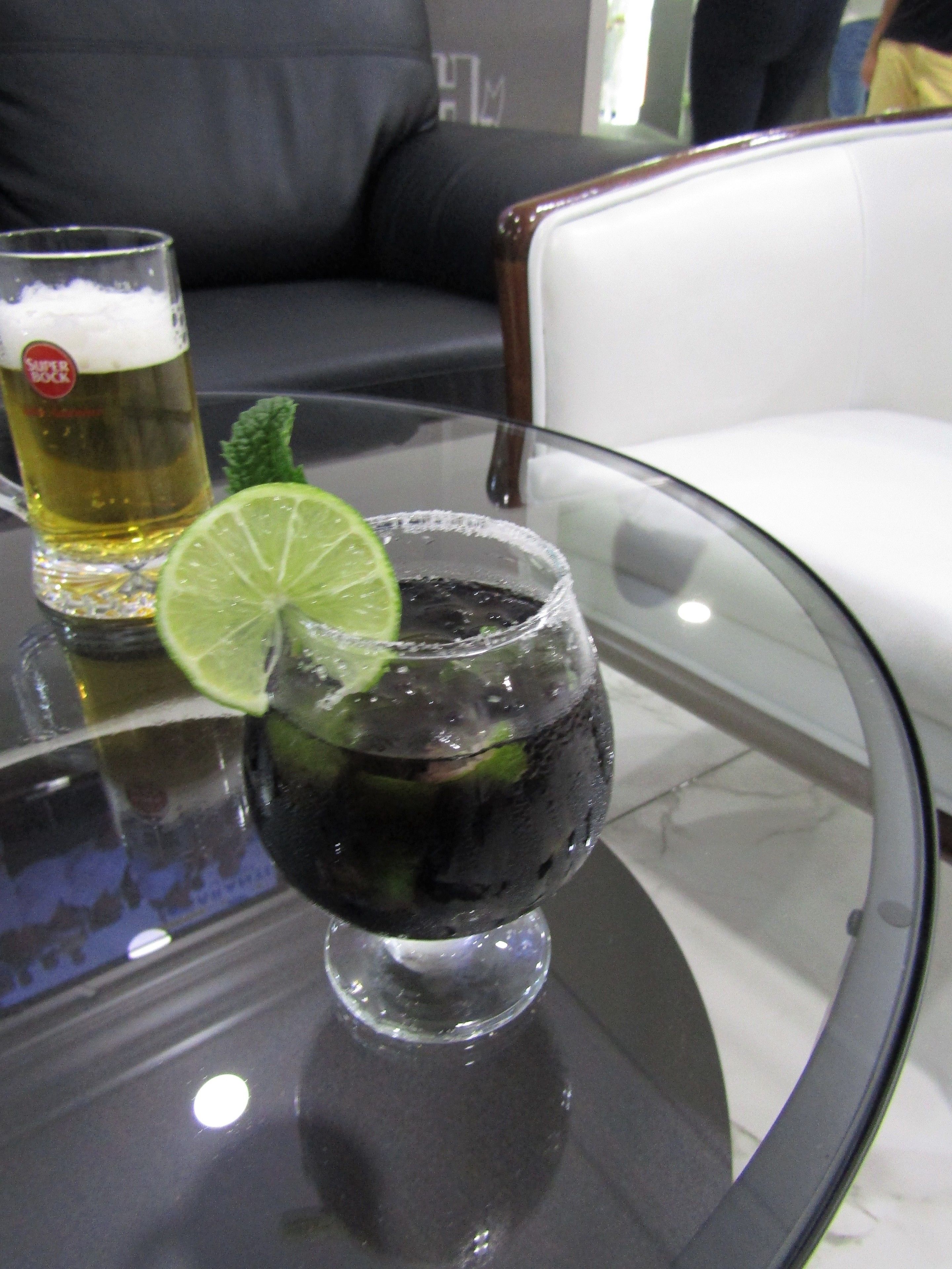A glass of beer with a lime slice on a glass table in a room with a black leather couch and a white chair.
