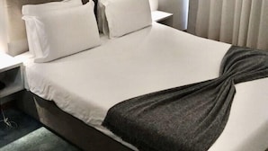 A hotel room with a large bed, white bedding, a headboard, a bedside table, a lamp, and a window with curtains.