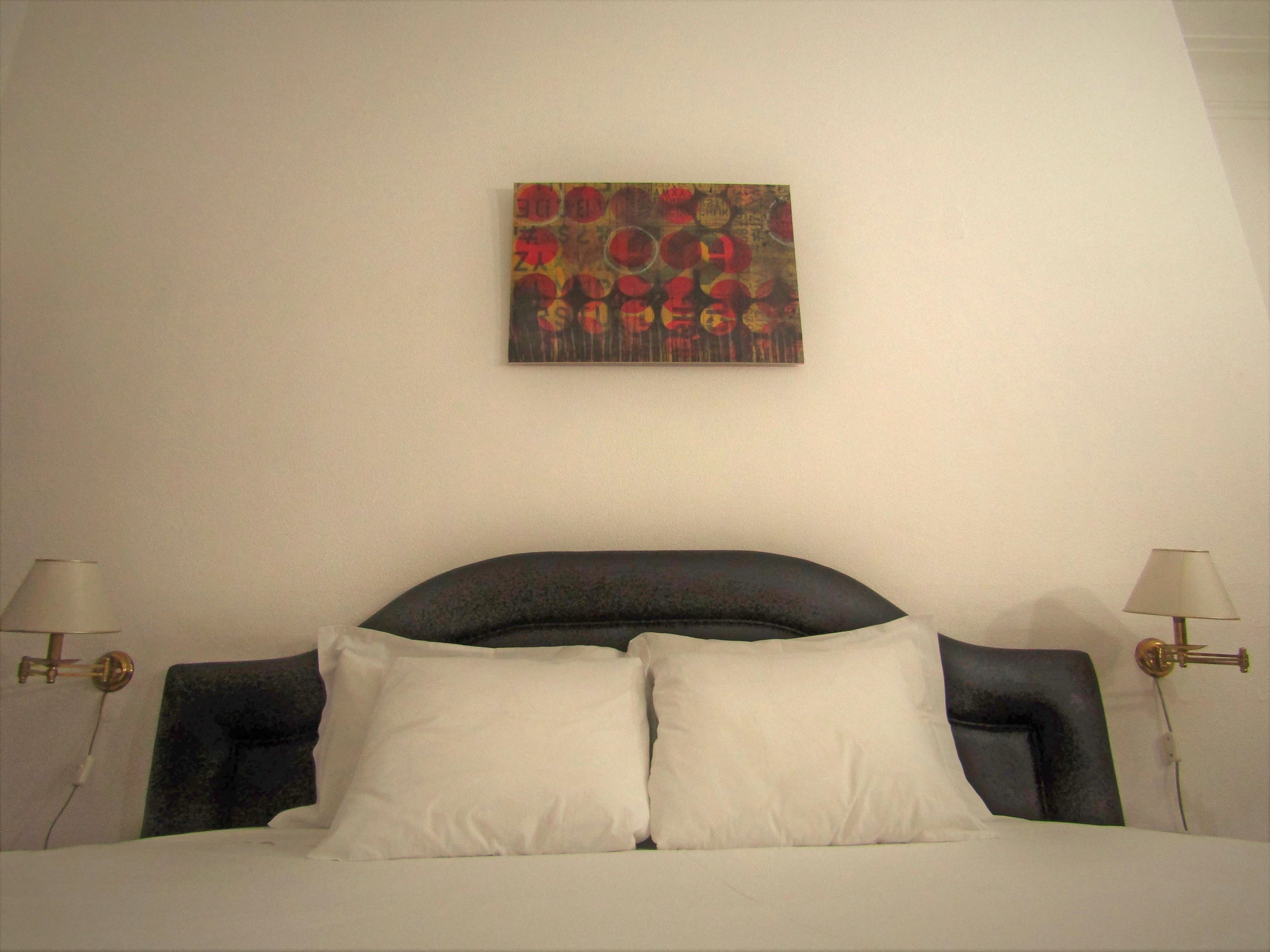 Ein Bett mit weißen Kissen und einem dunklen Kopfteil, das von zwei Nachttischlampen beleuchtet wird. Über dem Bett hängt ein farbenfrohes abstraktes Gemälde.