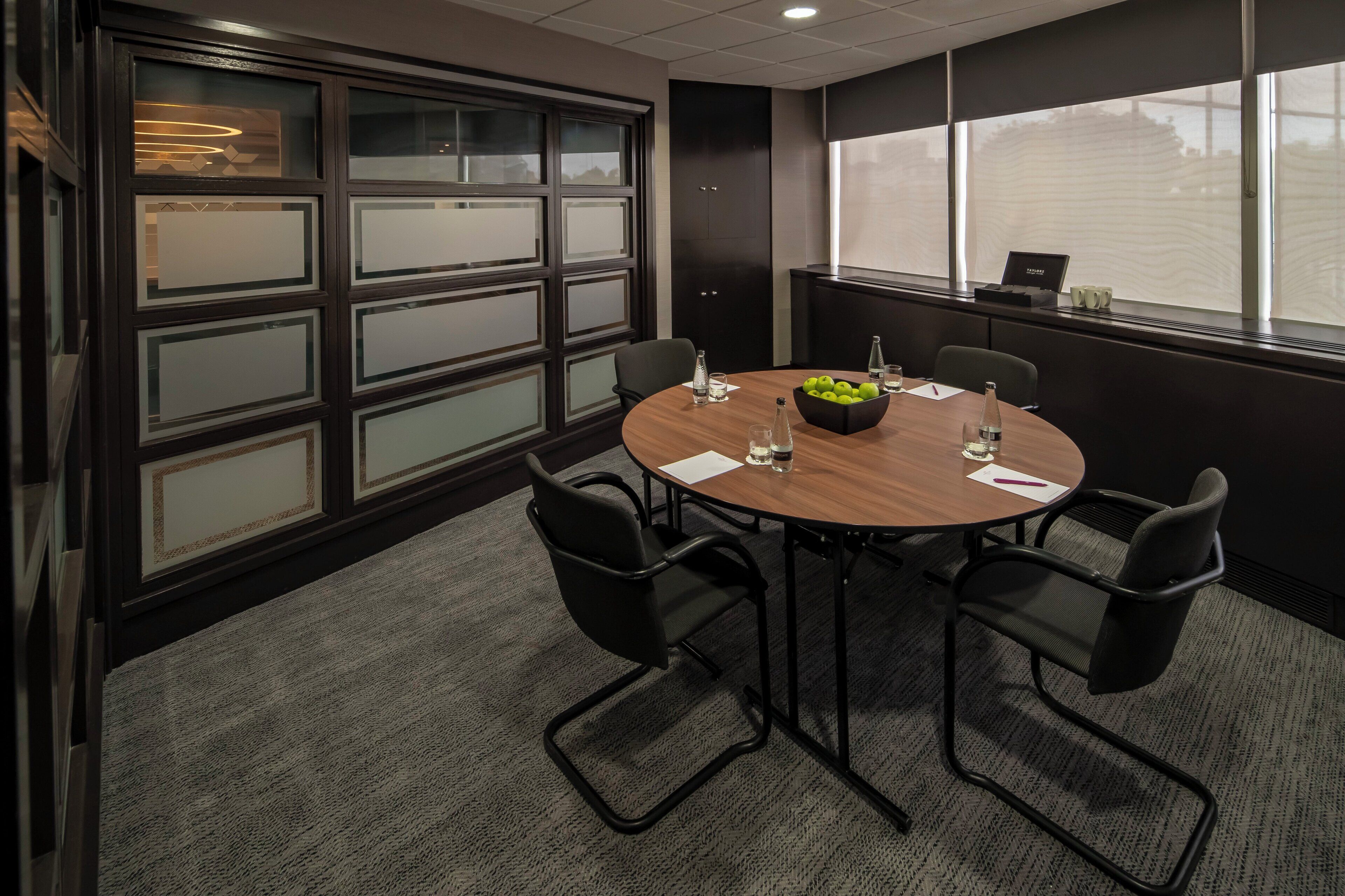 A conference room with a round table, four chairs, a bowl of fruit, and a window with blinds.