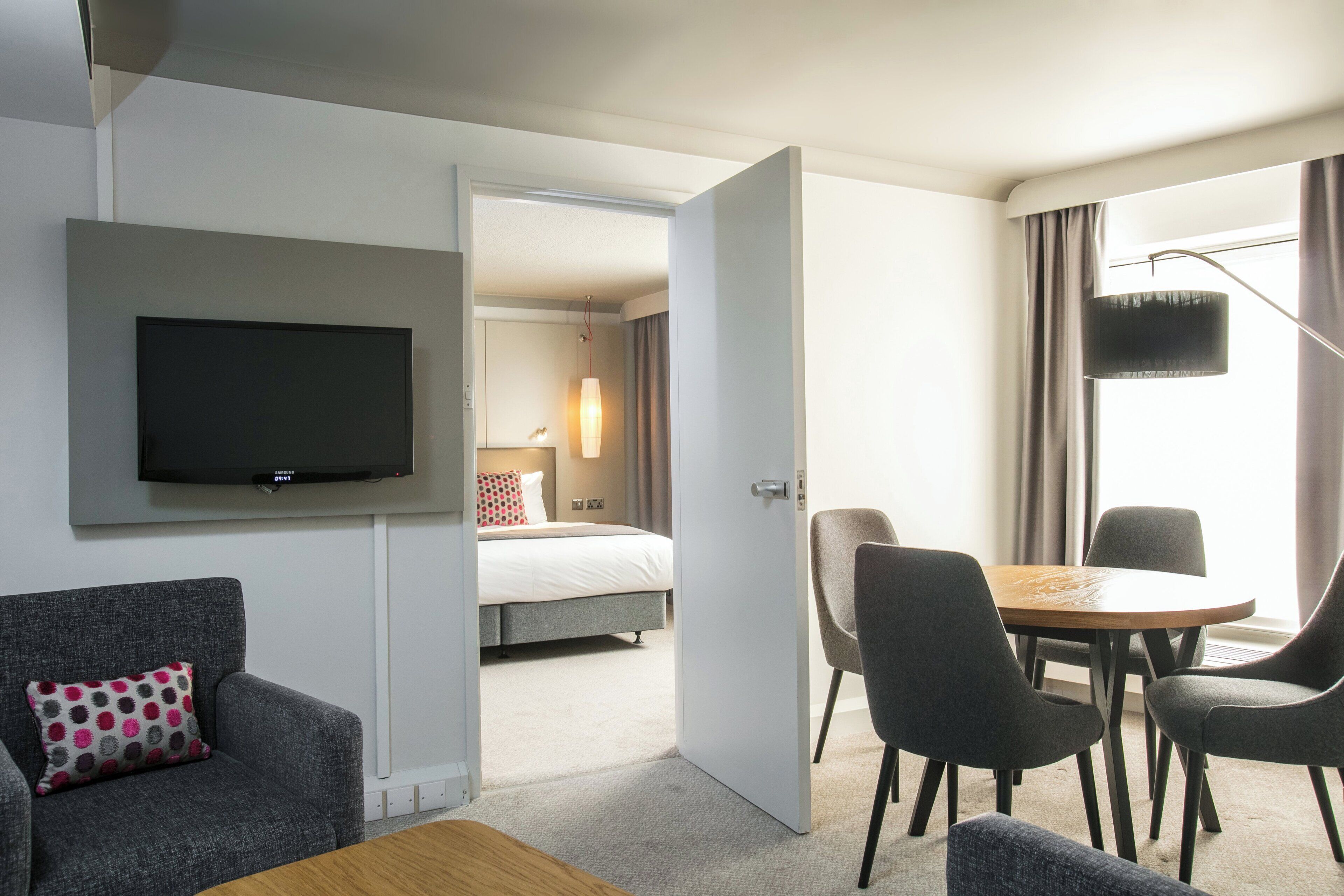 A hotel room with a flat-screen TV, a bed, a dining table with chairs, and a sofa with a patterned pillow.