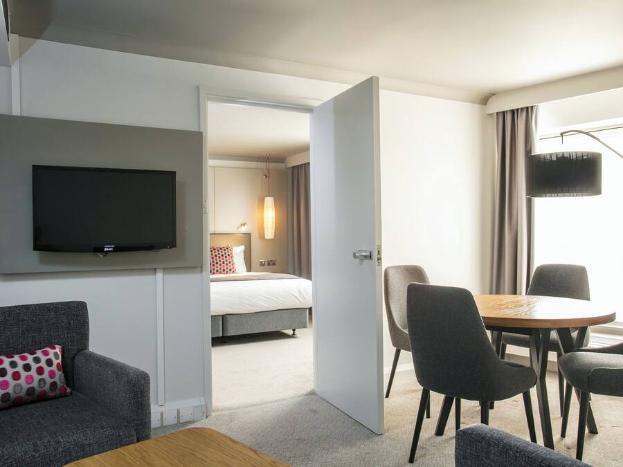 A hotel room with a flat-screen TV, a bed, a dining table with chairs, and a sofa with a patterned pillow.