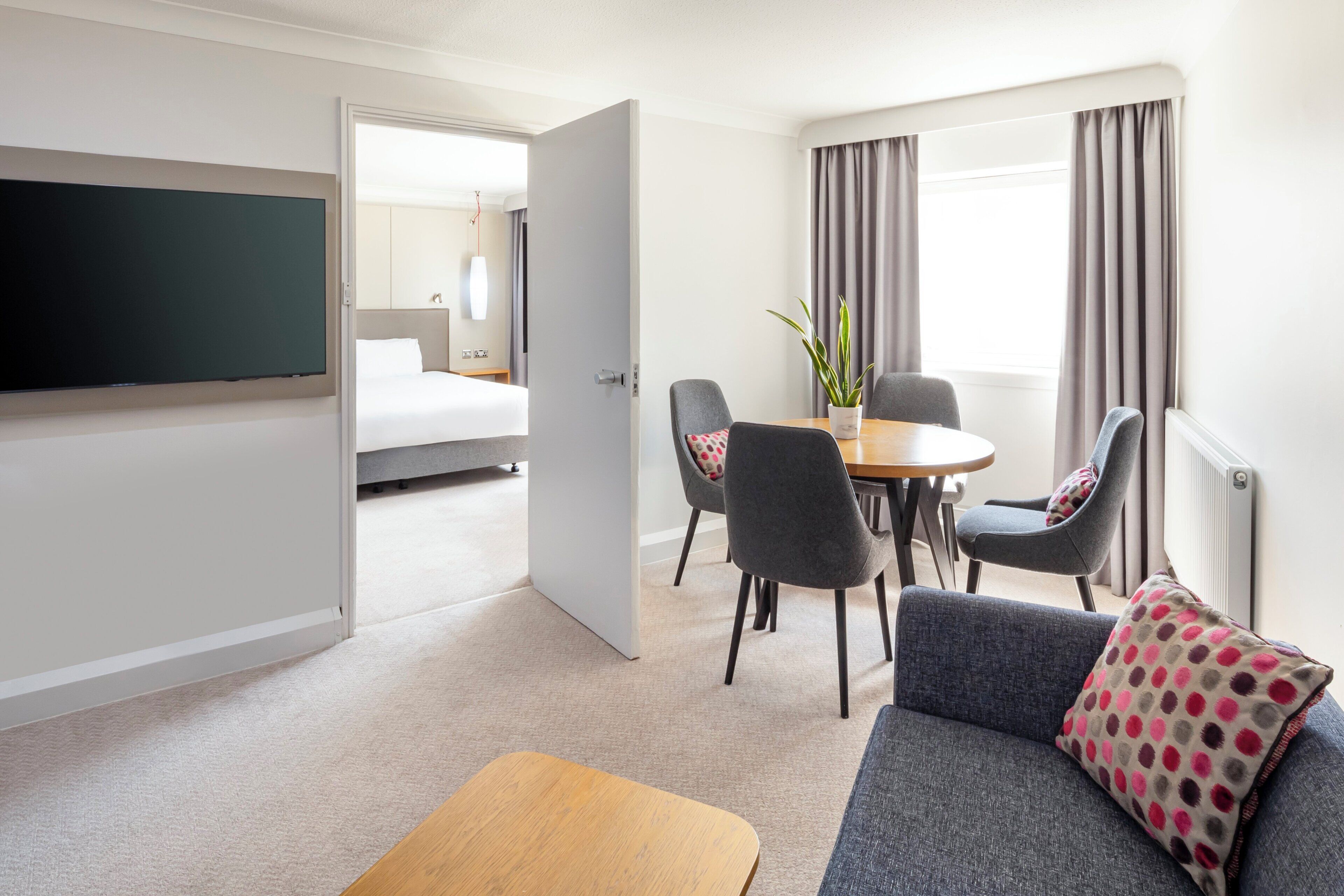 Een hotelkamer met een bed, een televisie, een eettafel met stoelen, een bank en een raam met gordijnen.