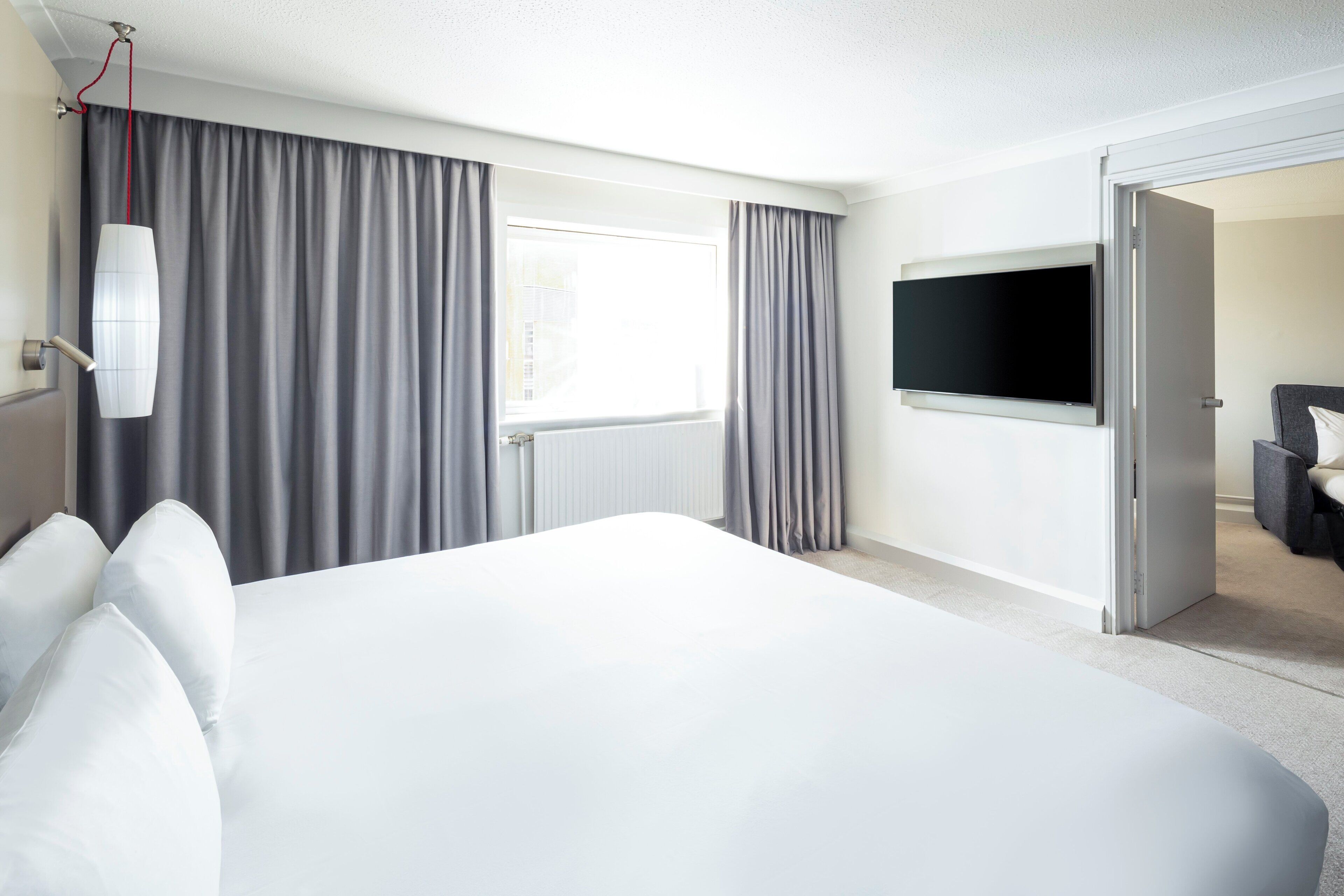 A hotel room with a large bed, a TV mounted on the wall, and a window with curtains.