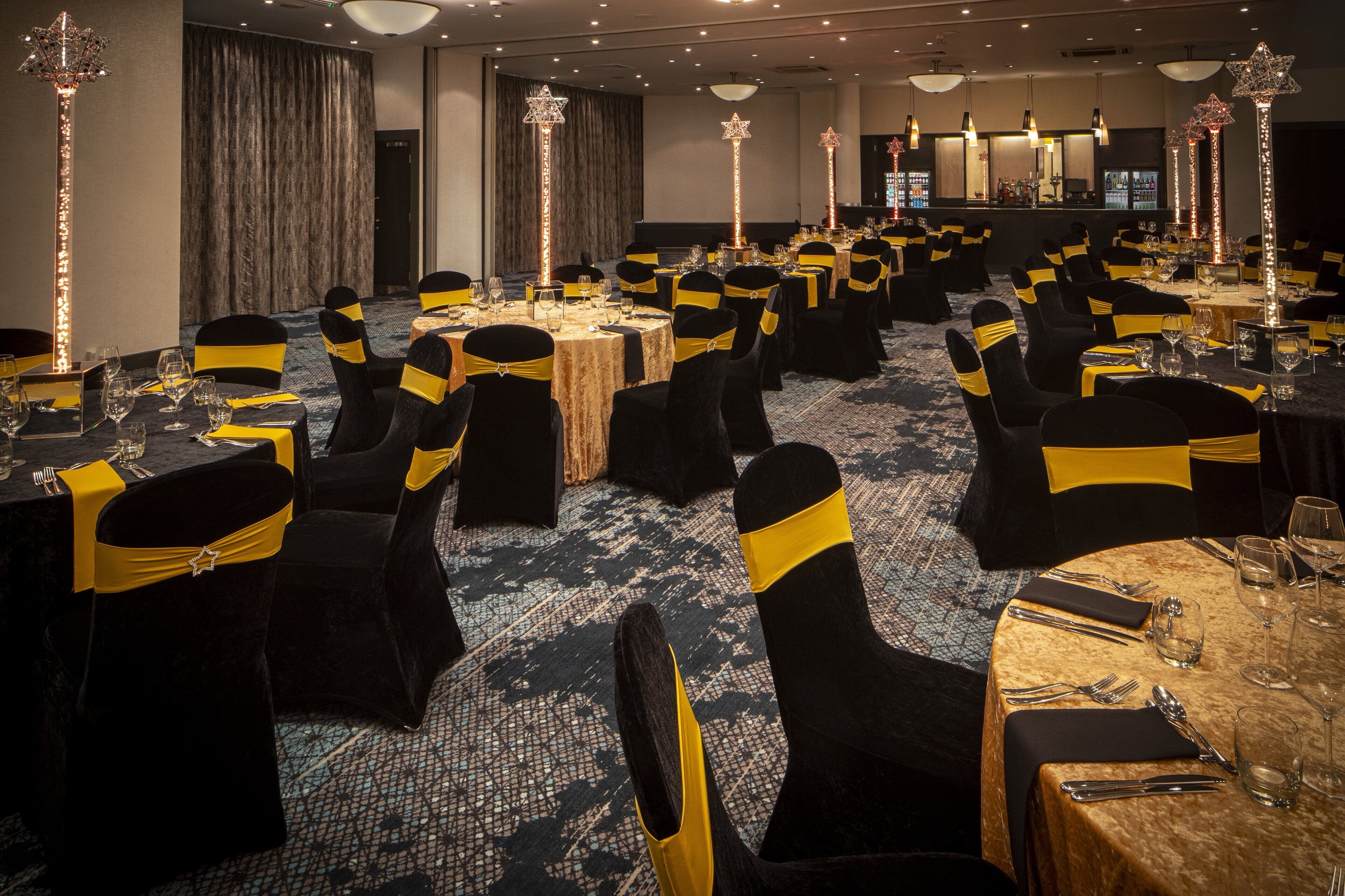 A banquet hall with round tables, black chairs with yellow sashes, and tall, star-shaped light fixtures.