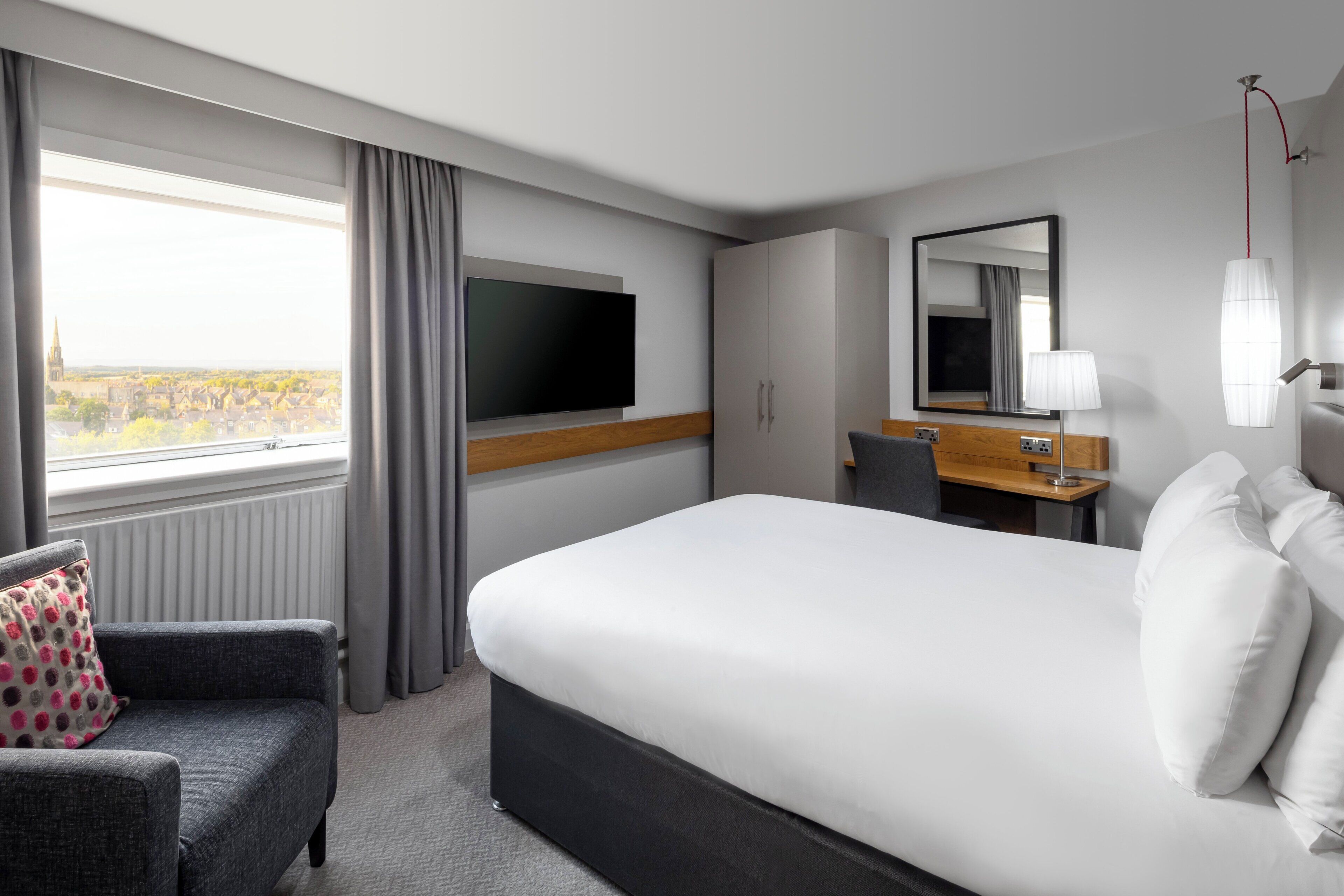 A hotel room with a bed, a TV, a desk, a chair, and a window with curtains.