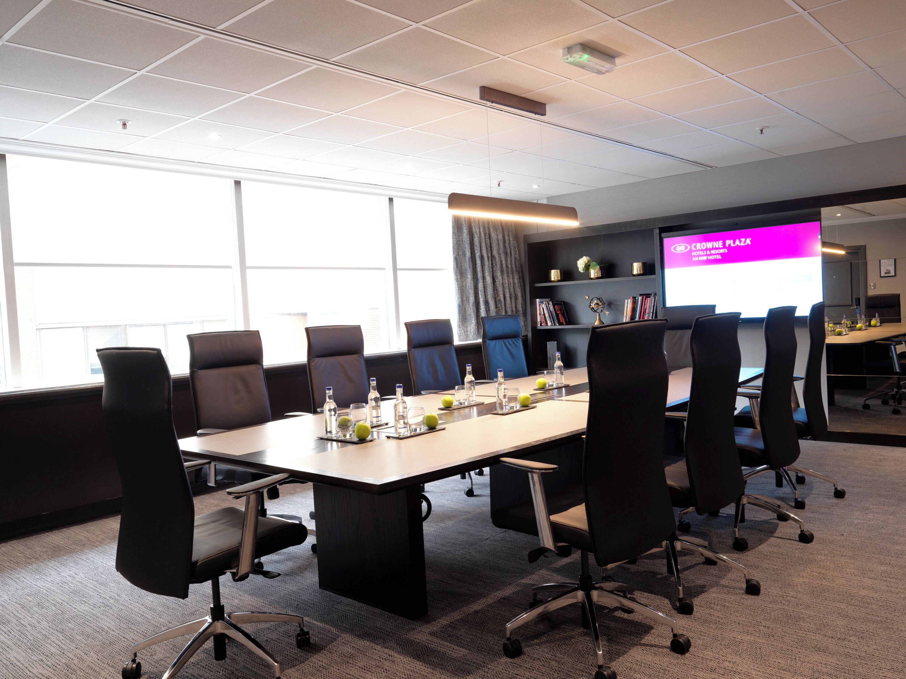 A conference room with a long table, chairs, a projector screen, and a bookshelf.