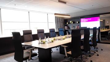 Un salón de conferencias con una larga mesa, sillas, una pantalla de proyector y una estantería.