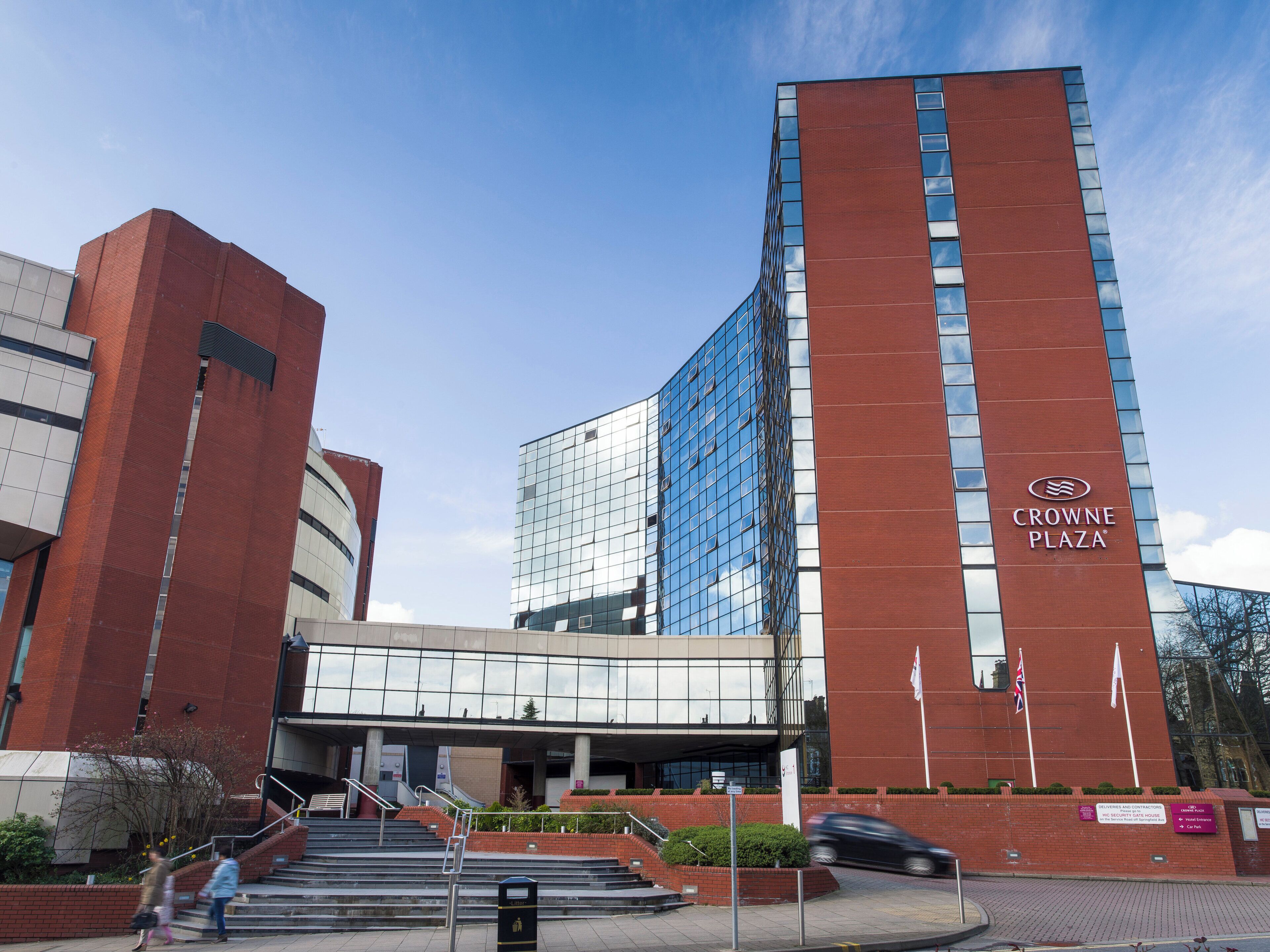 A modern building with a prominent 'CROWNE PLAZA' sign, featuring a mix of red brick and glass facades, and a set of stairs leading to the entrance.