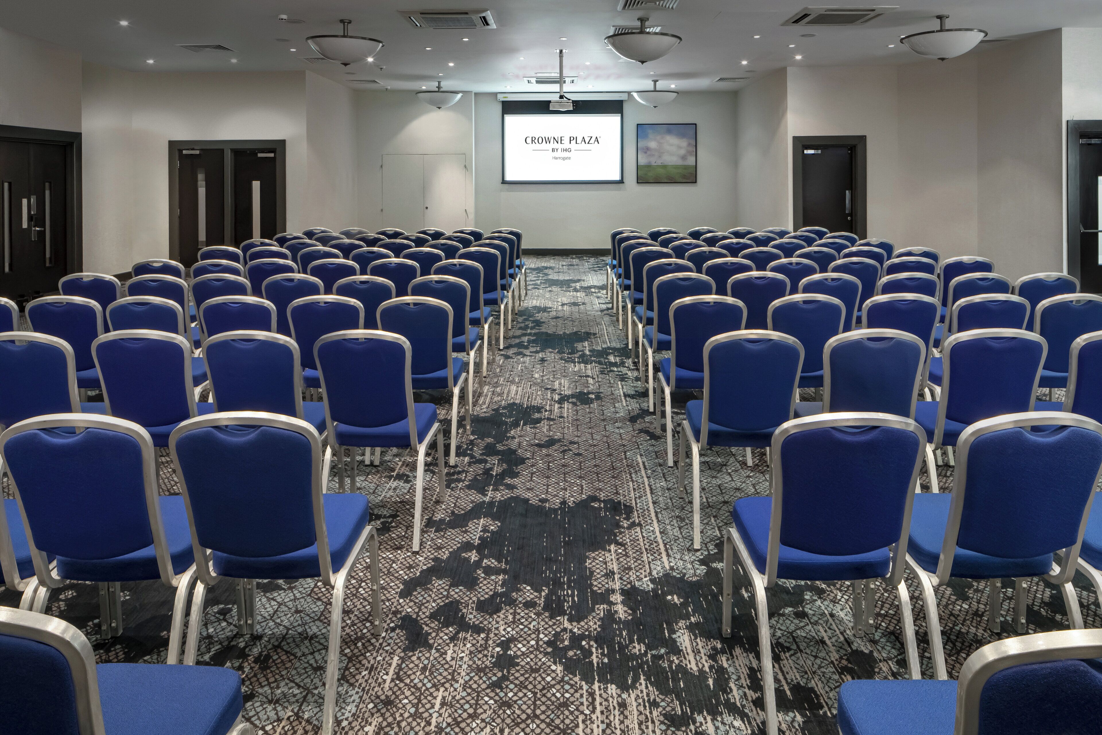Un salón de conferencias con filas de sillas azules, piso alfombrado, una pantalla de proyector y una puerta.
