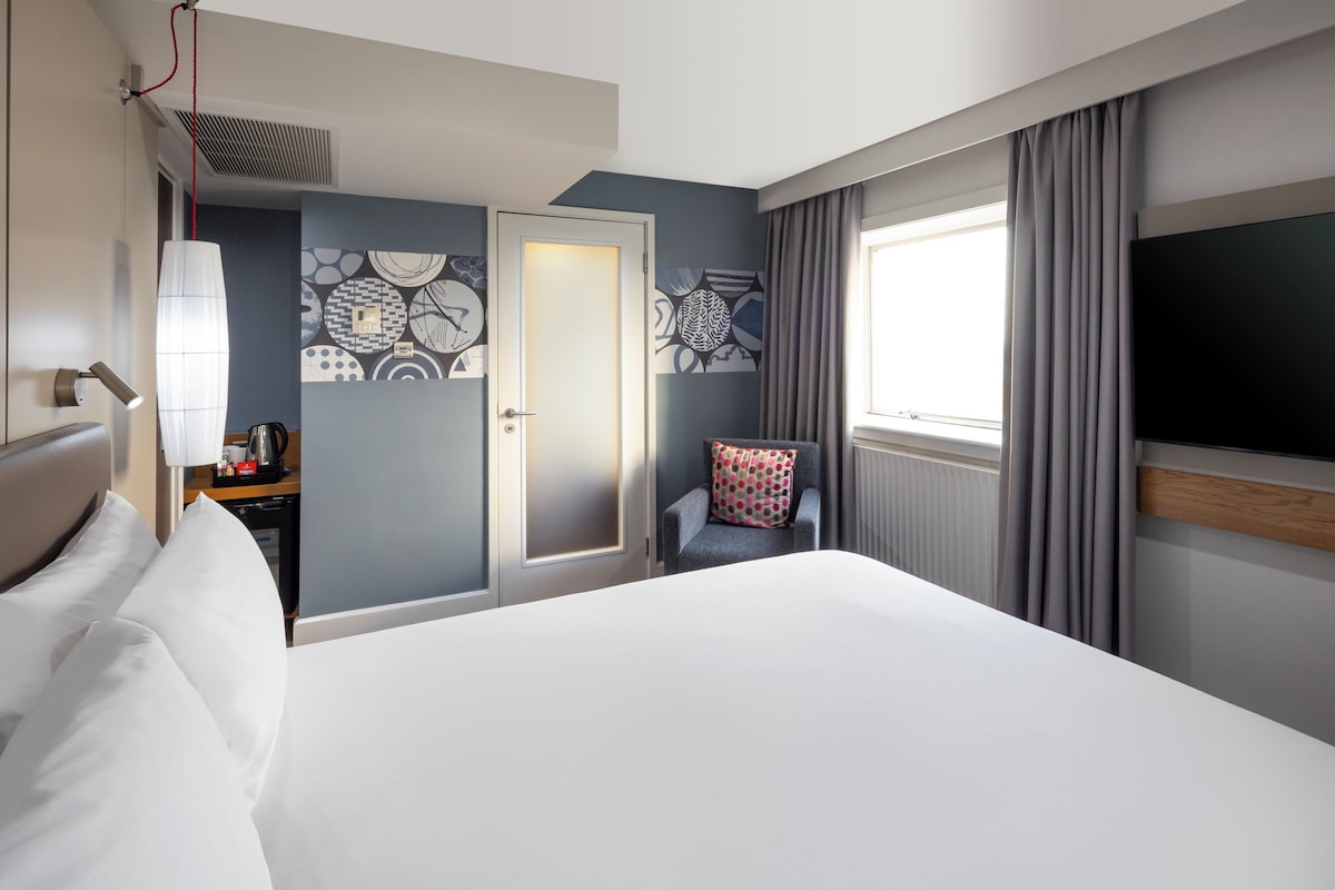 A hotel room with a large bed, a chair with a patterned cushion, a television, and a door leading to a bathroom.