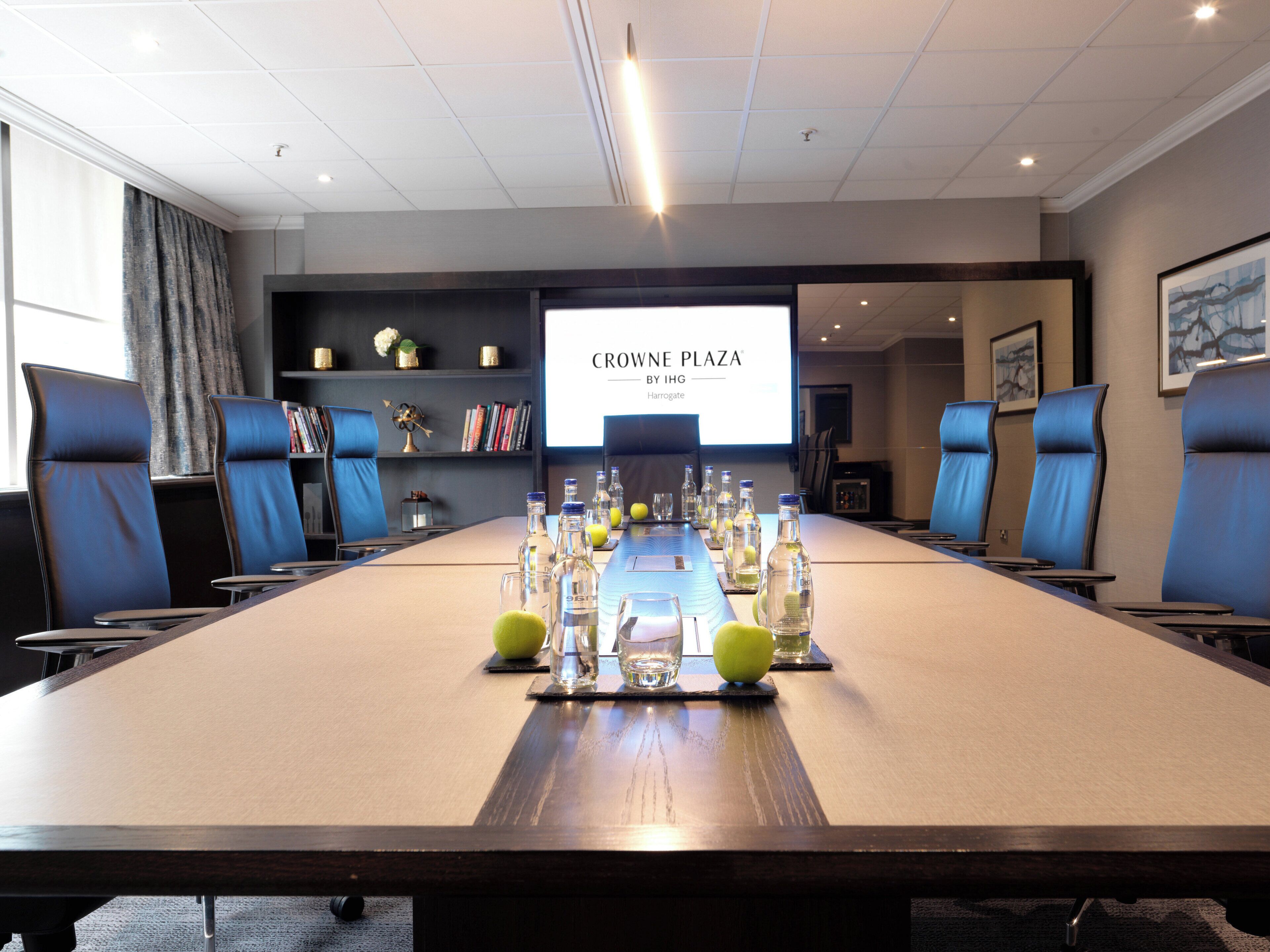 A conference room with a long table, blue chairs, a projector screen, and a bookshelf.