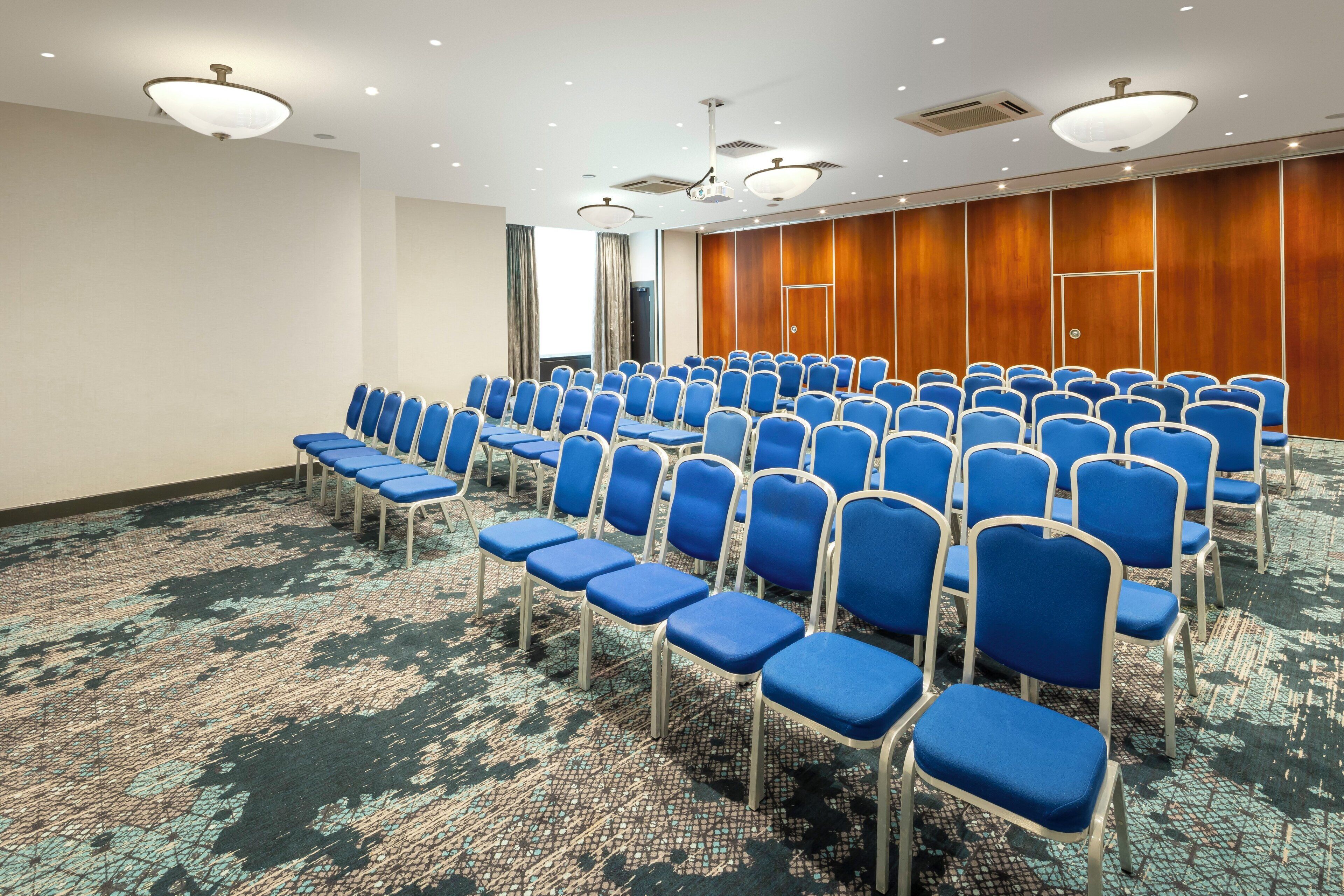 Une salle de conférence avec des rangées de chaises bleues, une moquette à motifs, des murs recouverts de panneaux en bois et des luminaires au plafond.