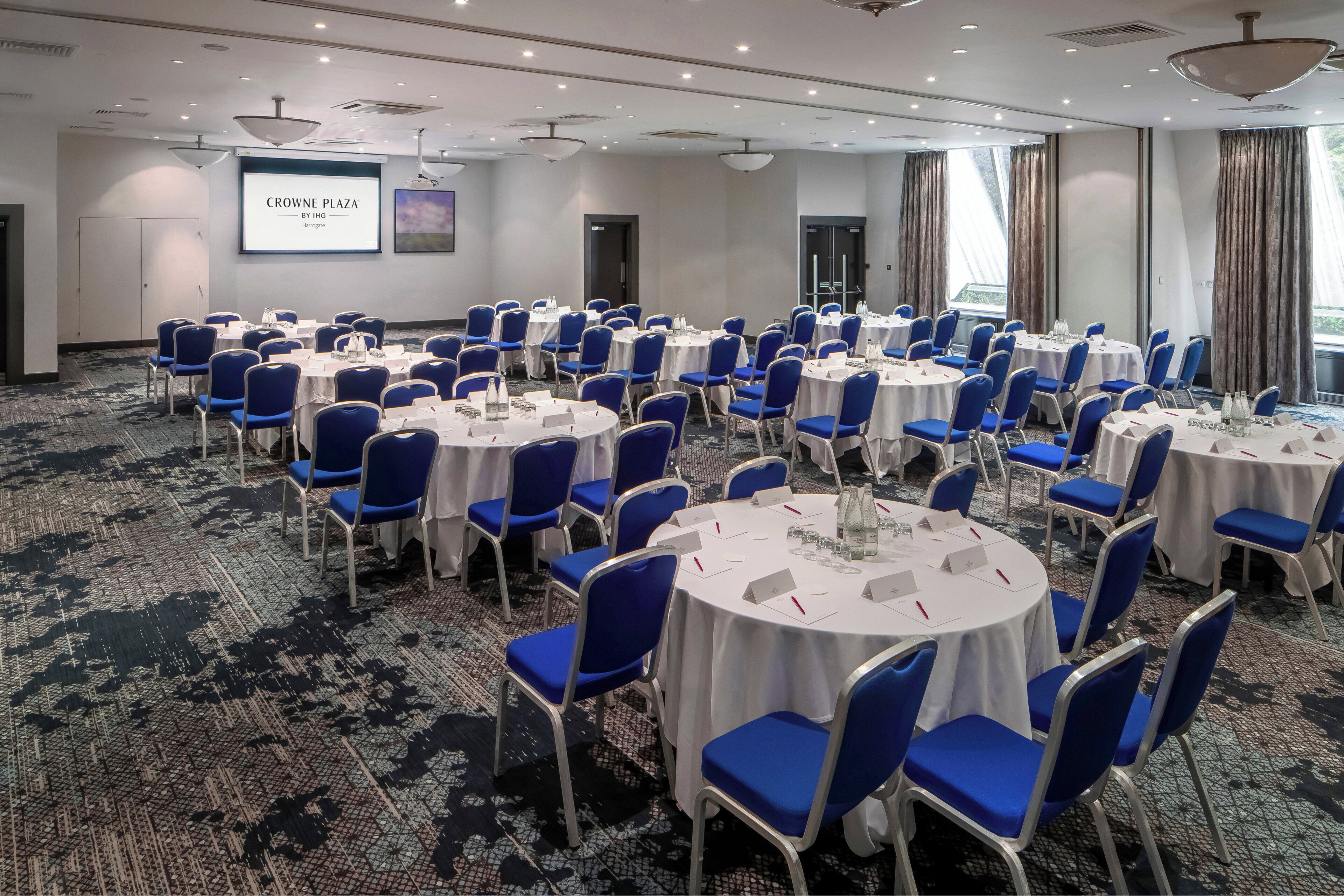 A conference room with round tables, blue chairs, and a projector screen displaying 'CROWNE PLAZA BY INO'.