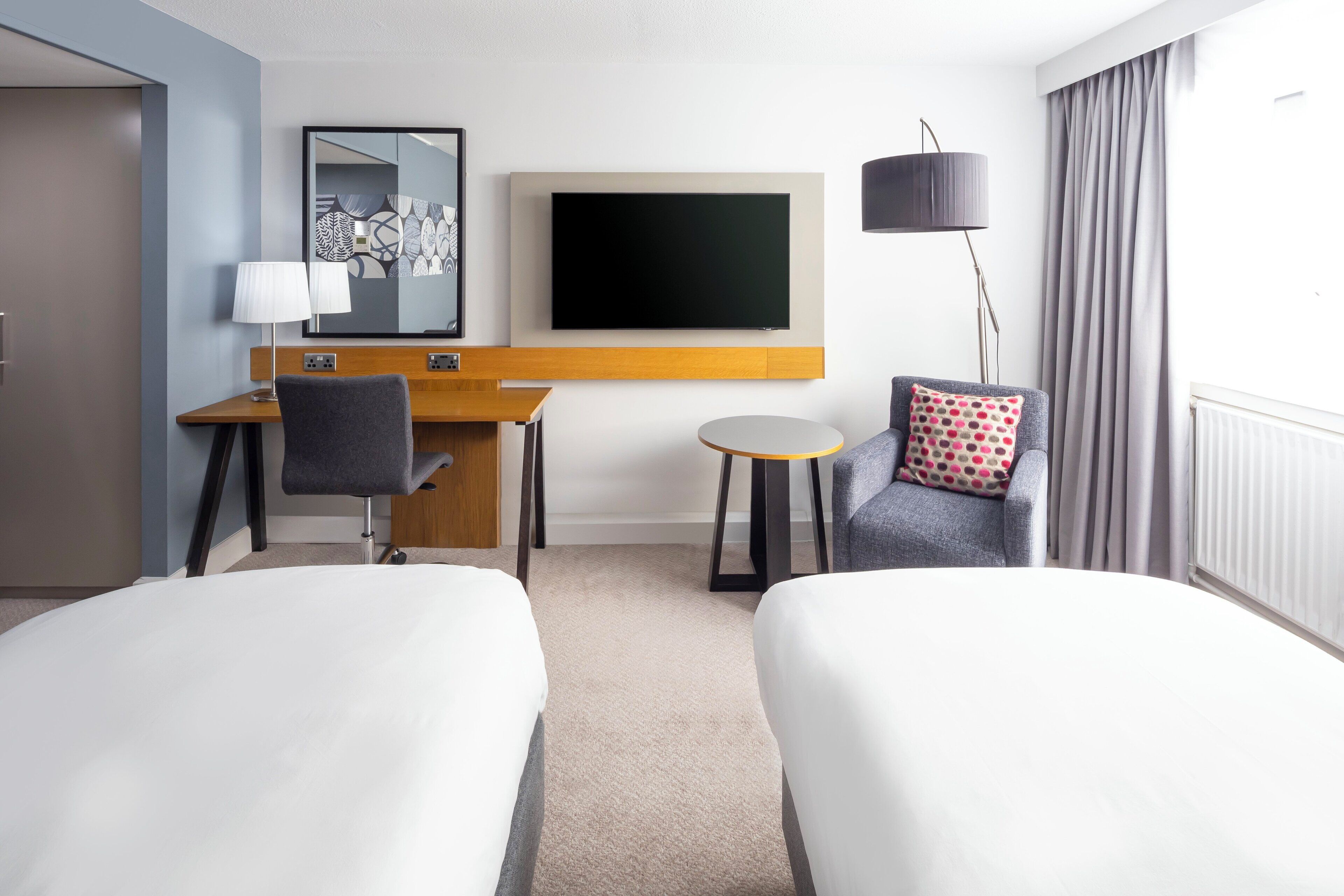 A hotel room with two beds, a desk, a chair, a TV, and a lamp.