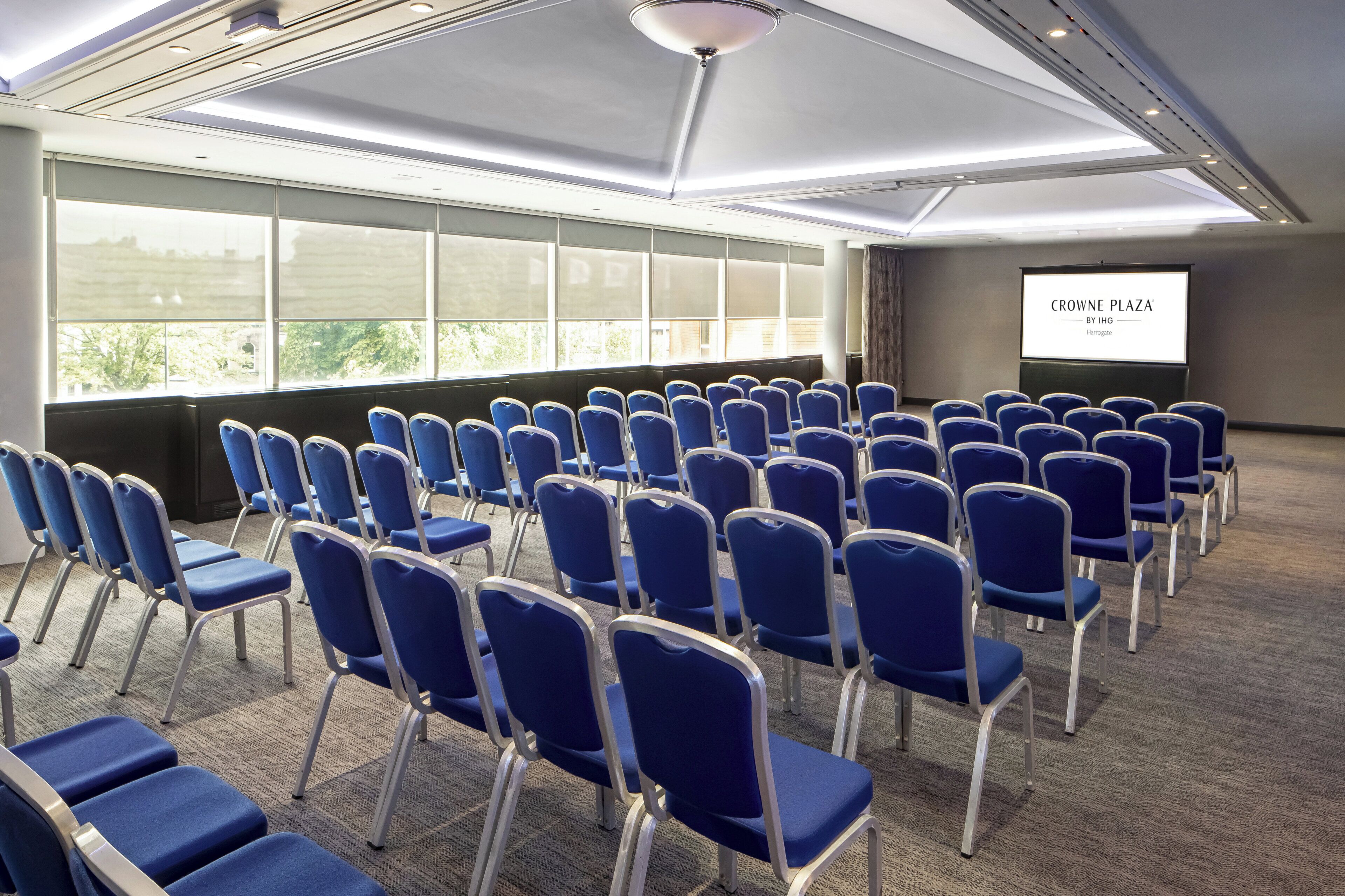 A conference room with rows of blue chairs, a large screen displaying 'CROWNE PLAZA BY IHG,' and large windows allowing natural light.