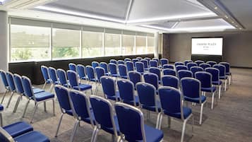A conference room with rows of blue chairs, a large screen displaying 'CROWNE PLAZA BY IHG,' and large windows allowing natural light.