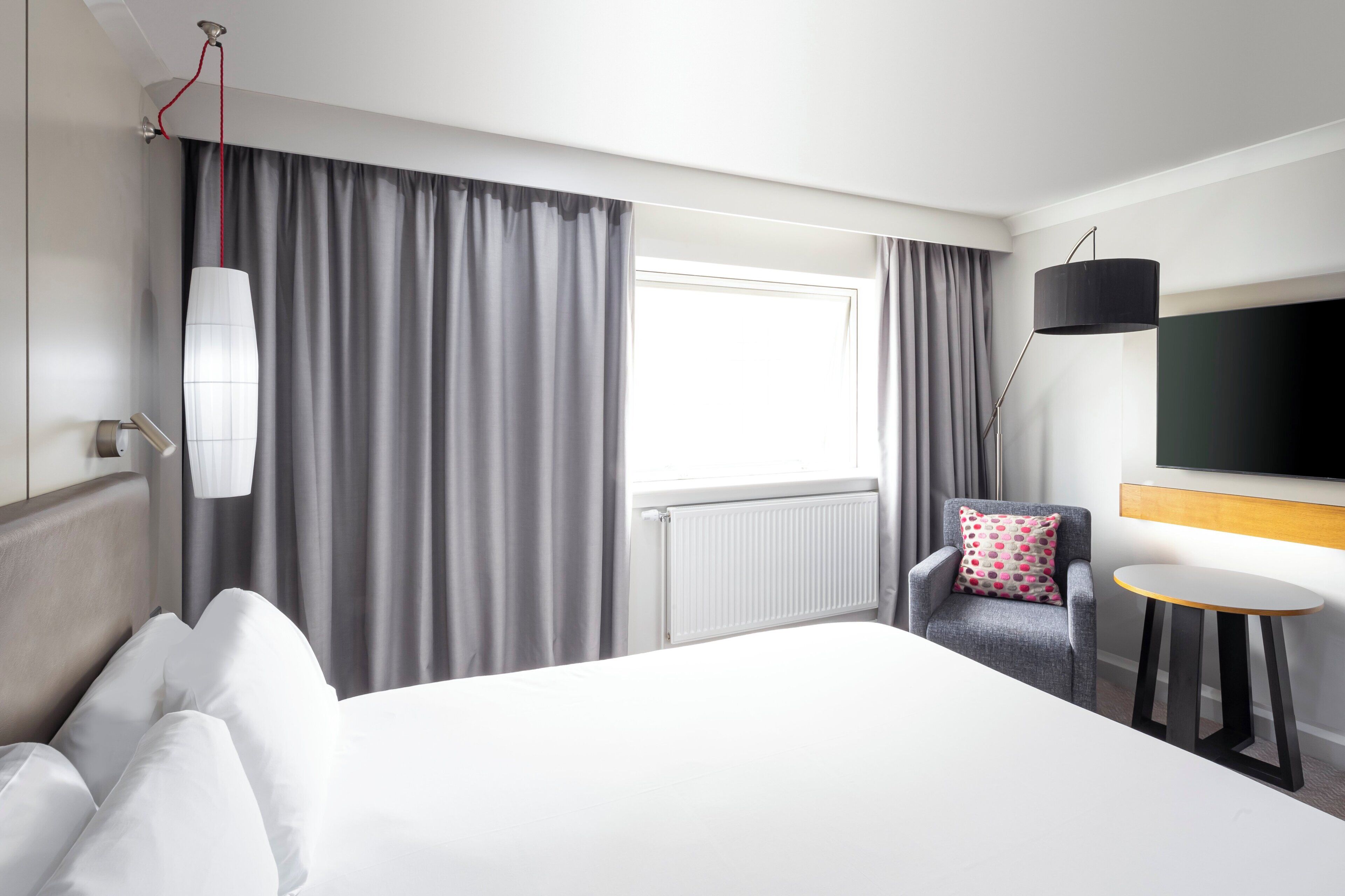 Une chambre d’hôtel avec un grand lit, une chaise, une petite table, un téléviseur et une fenêtre avec des rideaux.