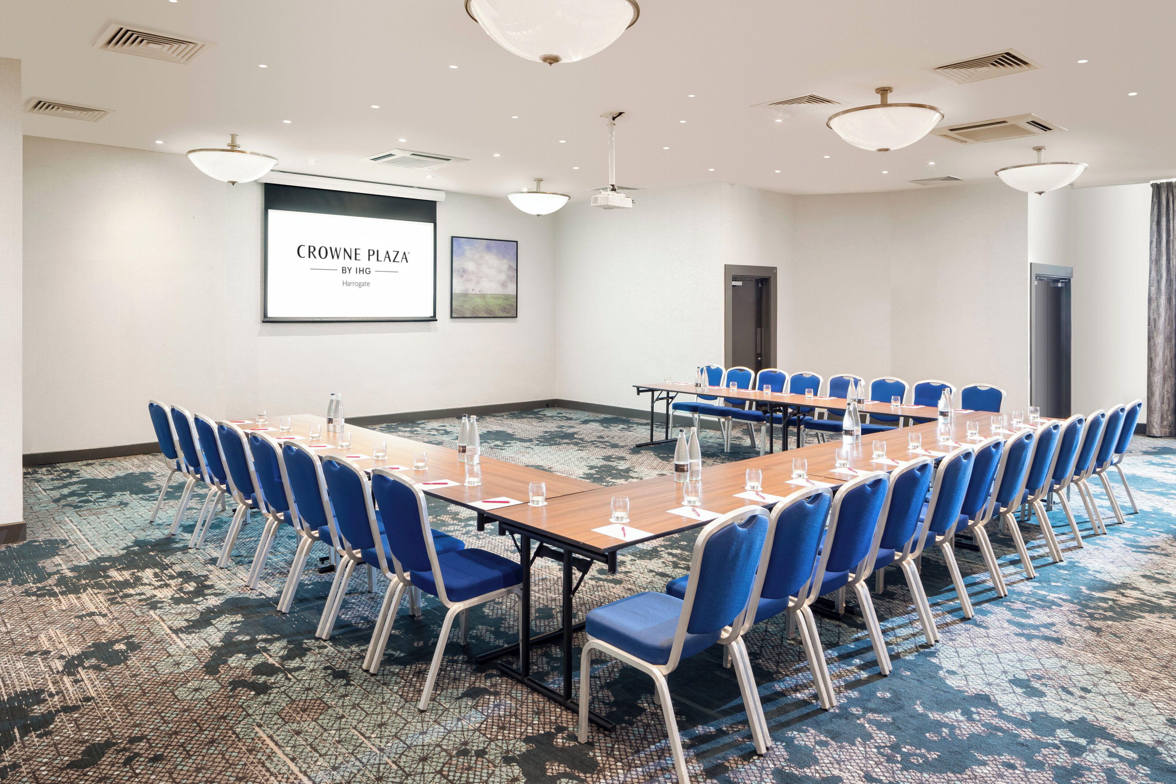 A conference room with a long table, blue chairs, a projector screen, and a carpeted floor.