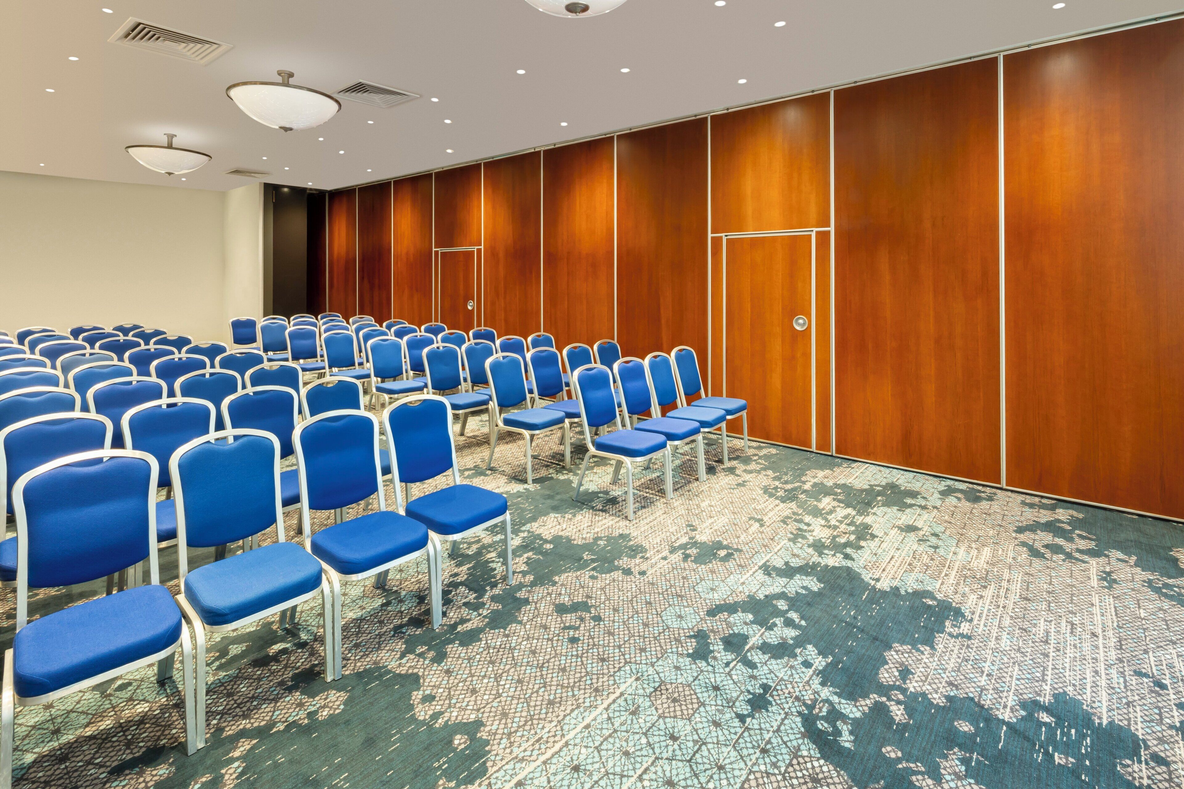 A conference room with rows of blue chairs, a patterned carpet, and wooden panel walls.