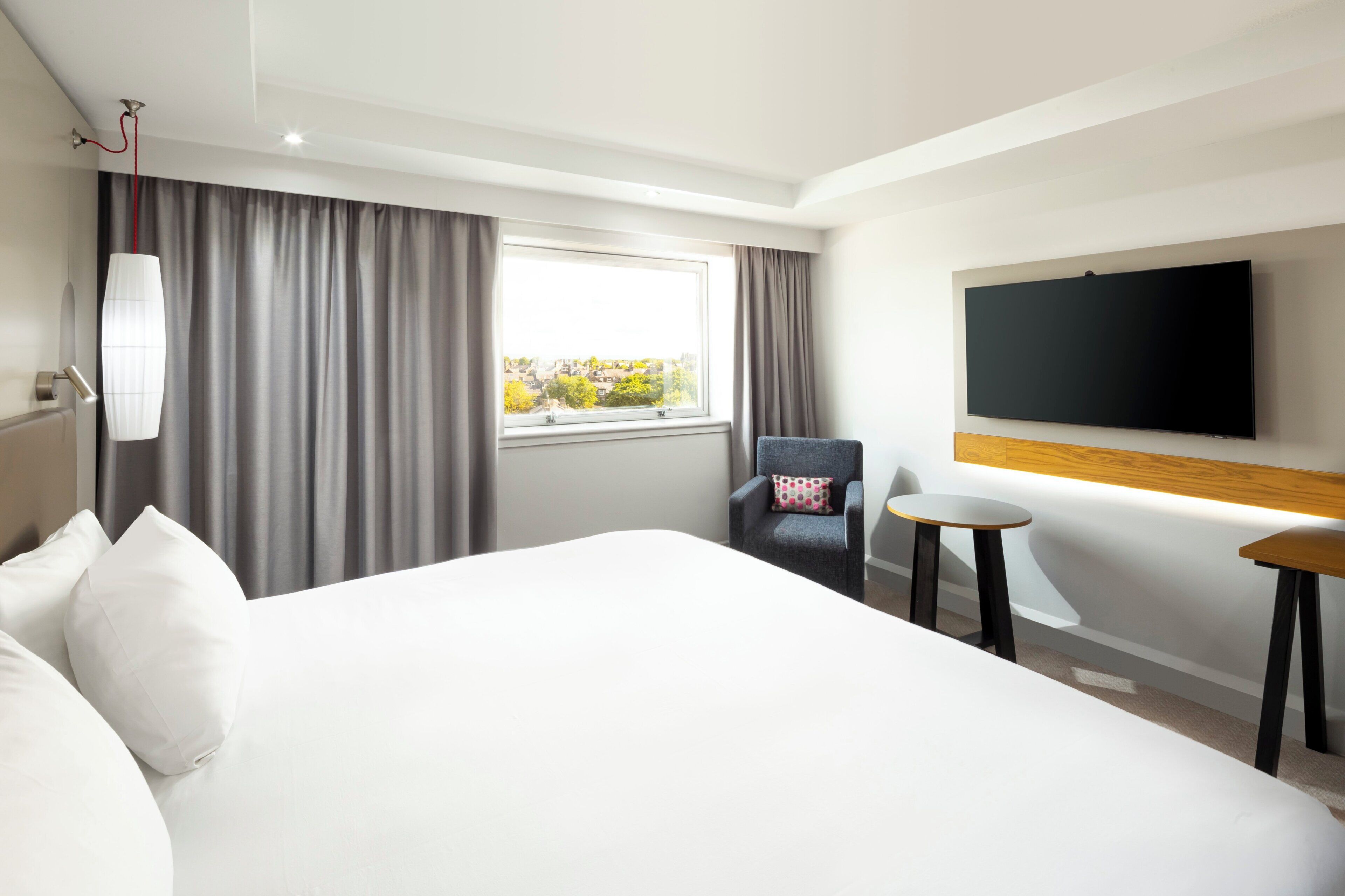 Une chambre d’hôtel avec un grand lit, une chaise, une petite table, un téléviseur et une fenêtre avec des rideaux.