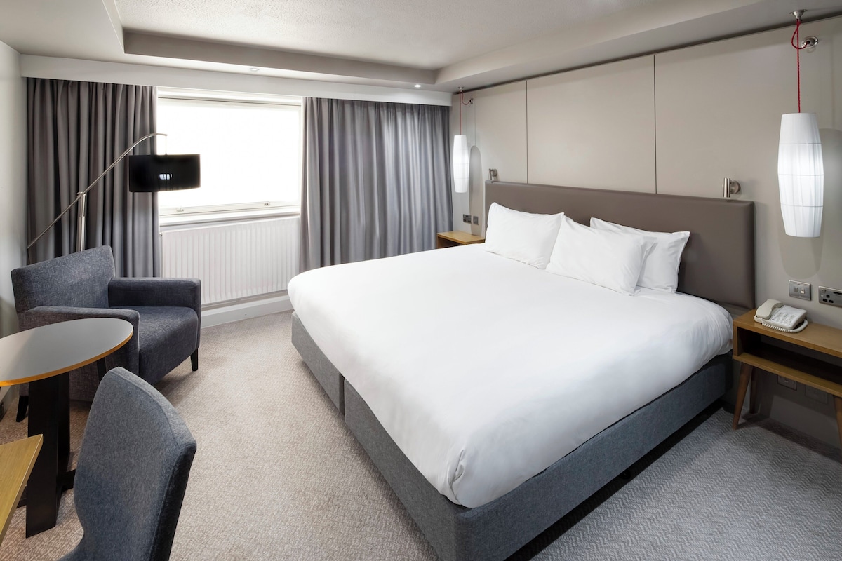 A hotel room with a large bed, a desk, a chair, a TV, and a window with curtains.