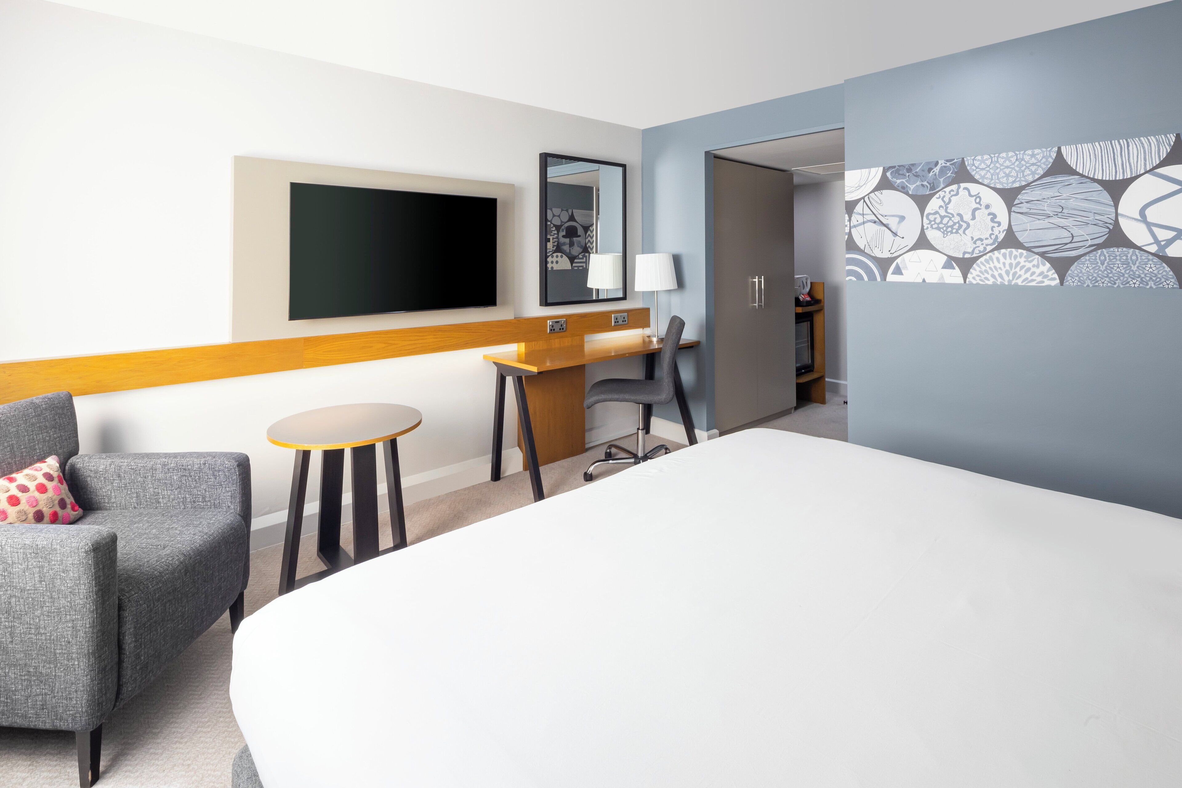 Ein modernes Hotelzimmer mit einem großen Bett, einem Schreibtisch mit Stuhl, einem kleinen runden Tisch, einer Couch und einem an der Wand befestigten Fernseher.