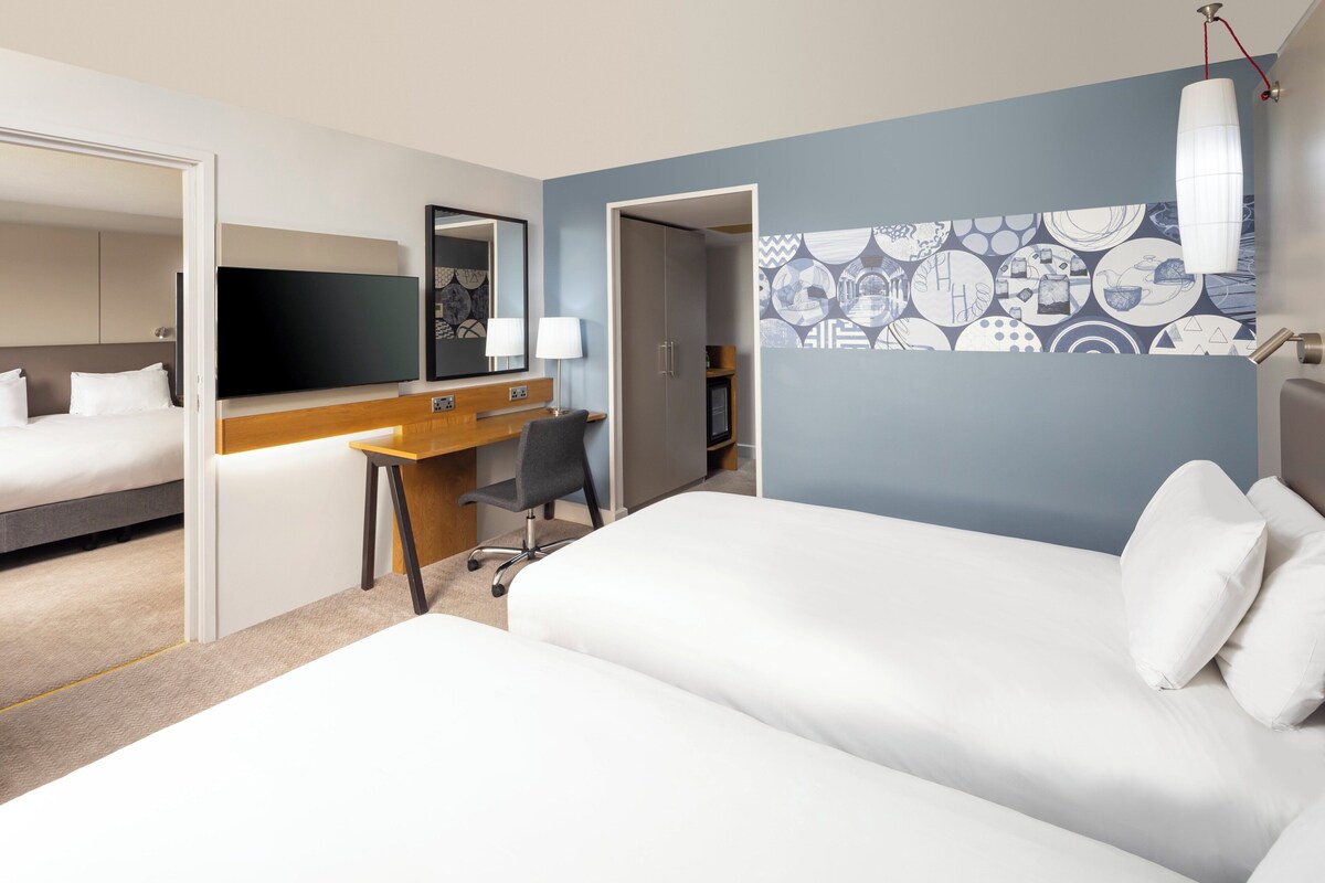 Habitación de hotel con cama, escritorio, silla, televisor y una puerta que da a otra habitación.