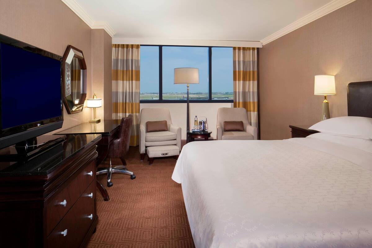 A hotel room with a large bed, a desk with a television, two armchairs, a dresser, and a large window with curtains.
