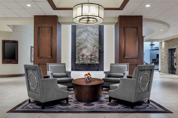 A lobby with a round wooden coffee table, four grey armchairs, a large chandelier, and a decorative wall with a chevron pattern. - Sheraton DFW Airport Hotel (Irving)