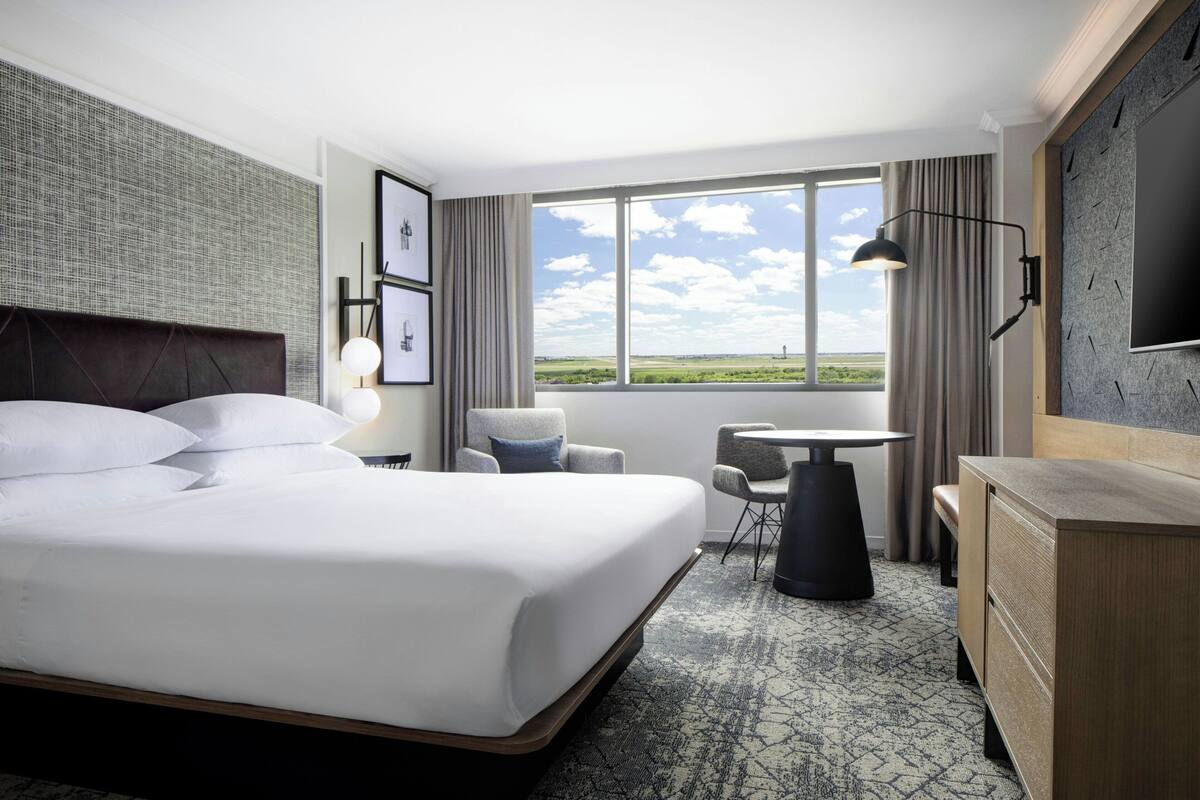 A hotel room with a large bed, a desk, a chair, a TV, and a window with a view of the countryside.