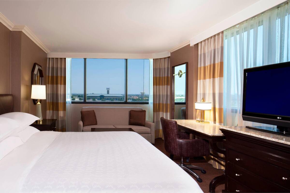 A hotel room with a large bed, a desk with a chair, a television, and a view of the cityscape.